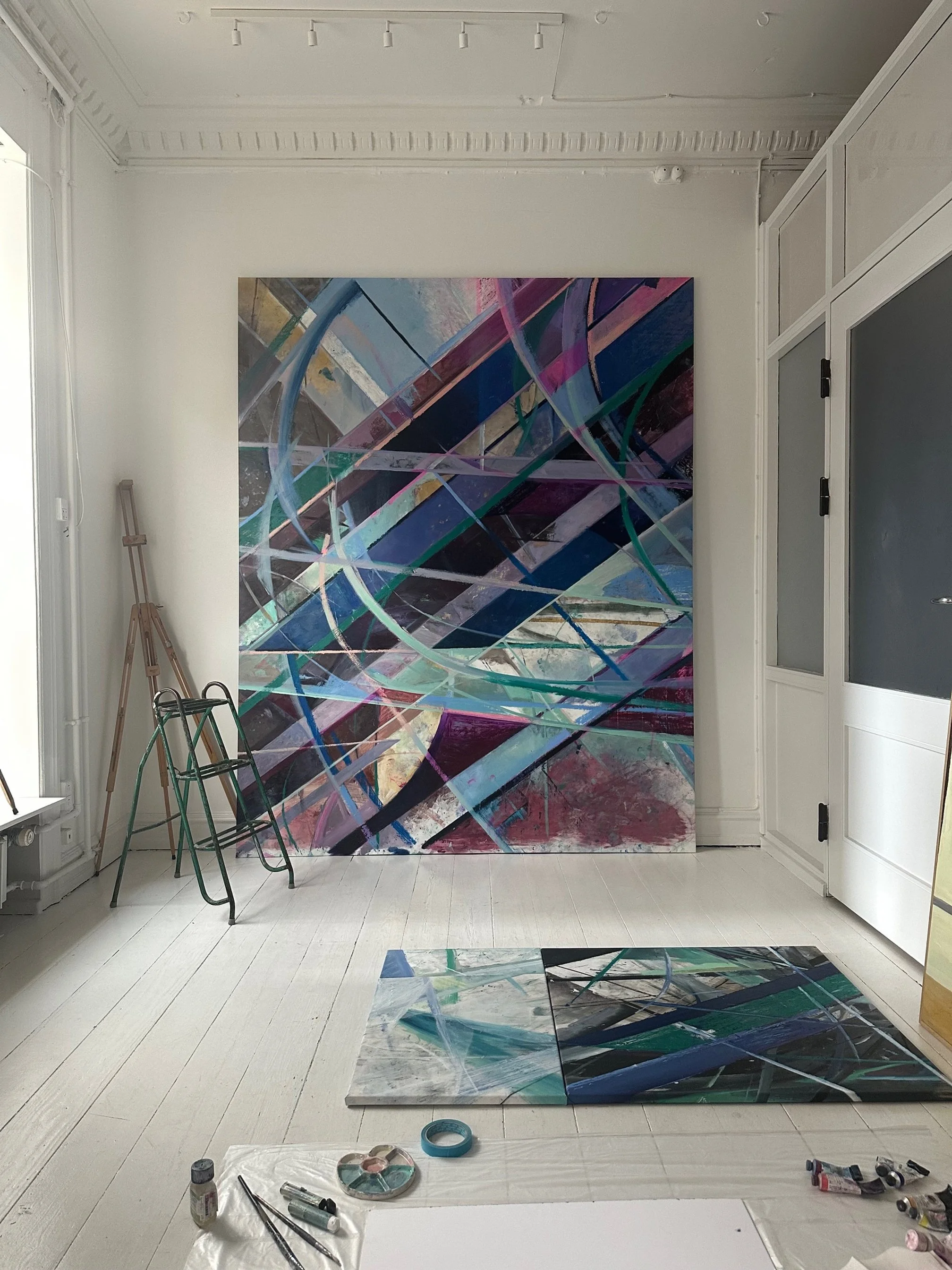 Abstract colorful painting with intersecting lines and geometric shapes resting against a white wall in an art studio, with painting supplies and a smaller artwork on the floor.