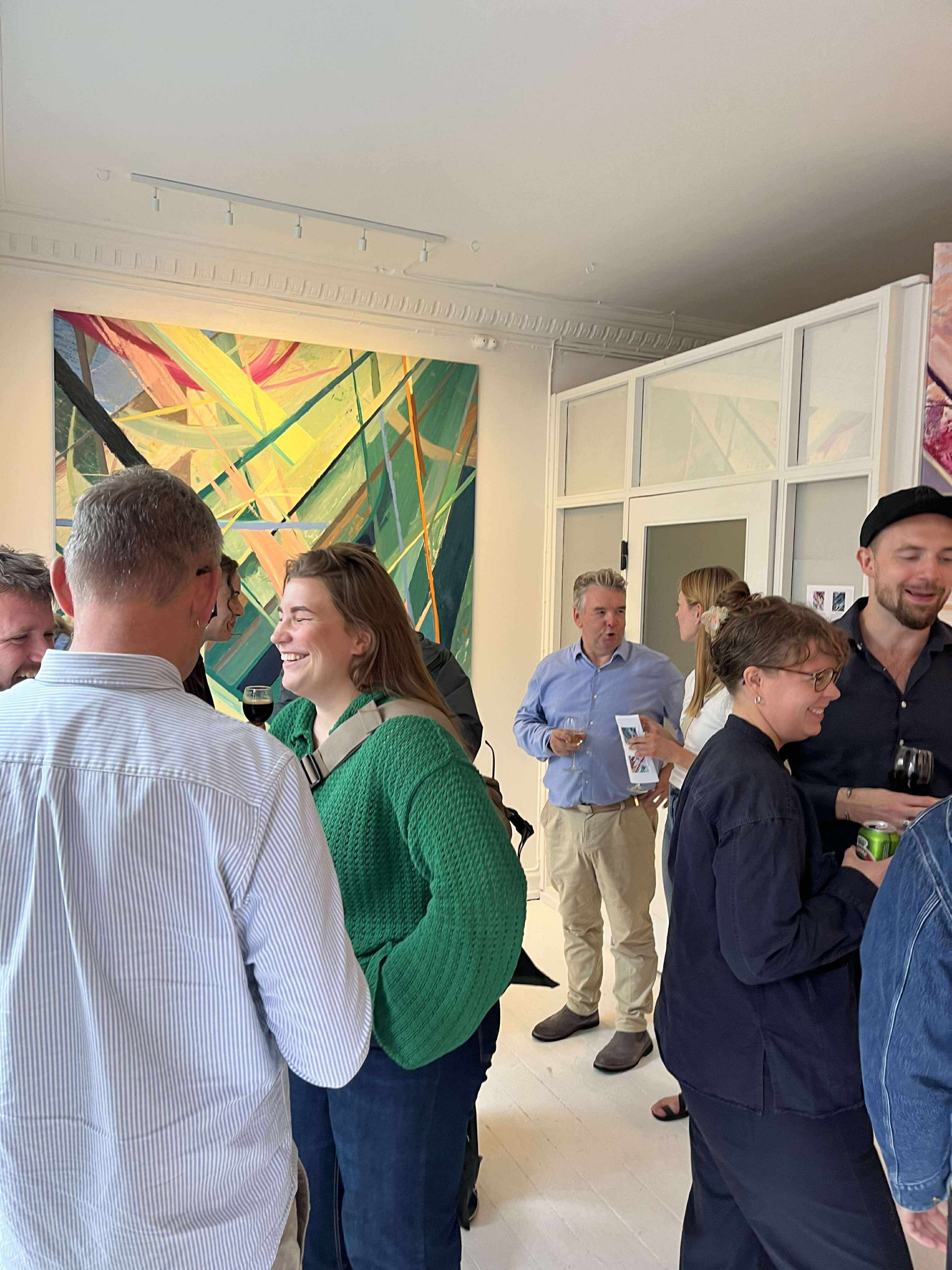 A group of people socializing at an art gallery, some holding drinks, with colorful abstract artwork on the wall behind them.