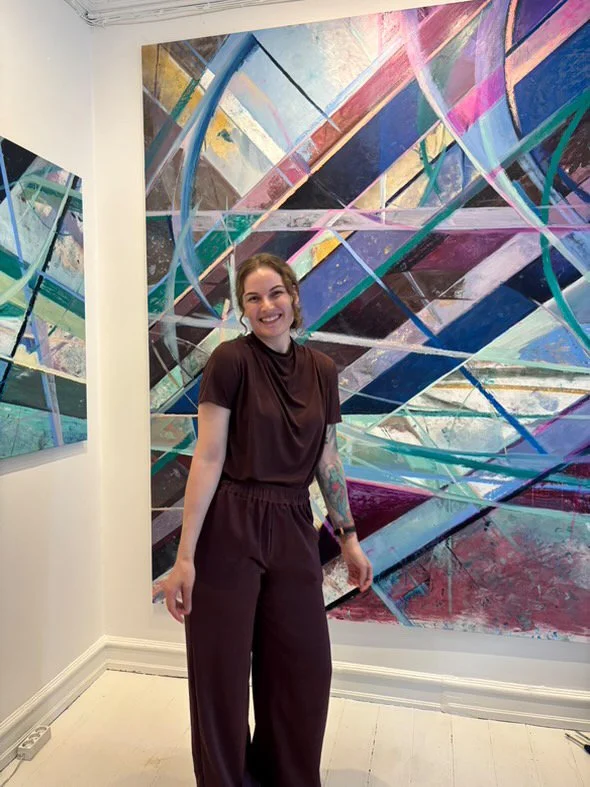 A woman with tattoos, wearing a dark brown outfit, smiling and posing in front of an abstract colorful artwork at an art gallery.