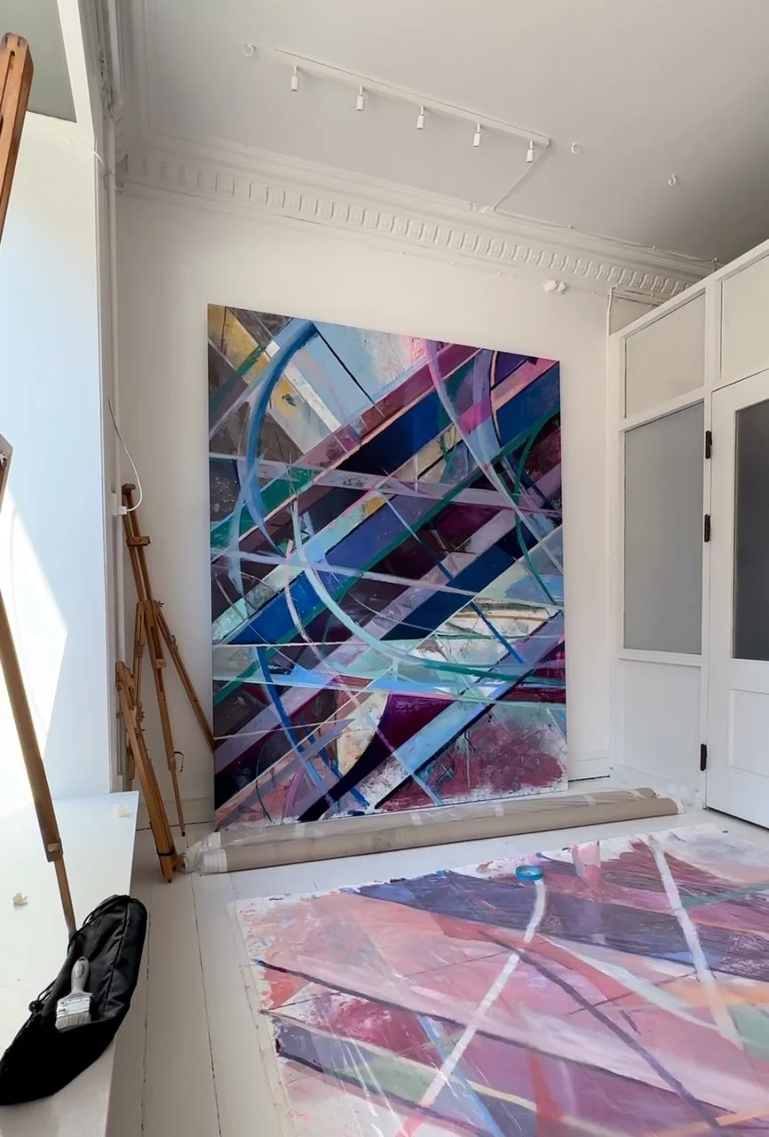 An abstract painting with intersecting colorful lines and geometric shapes leaning against a white wall in an art studio.