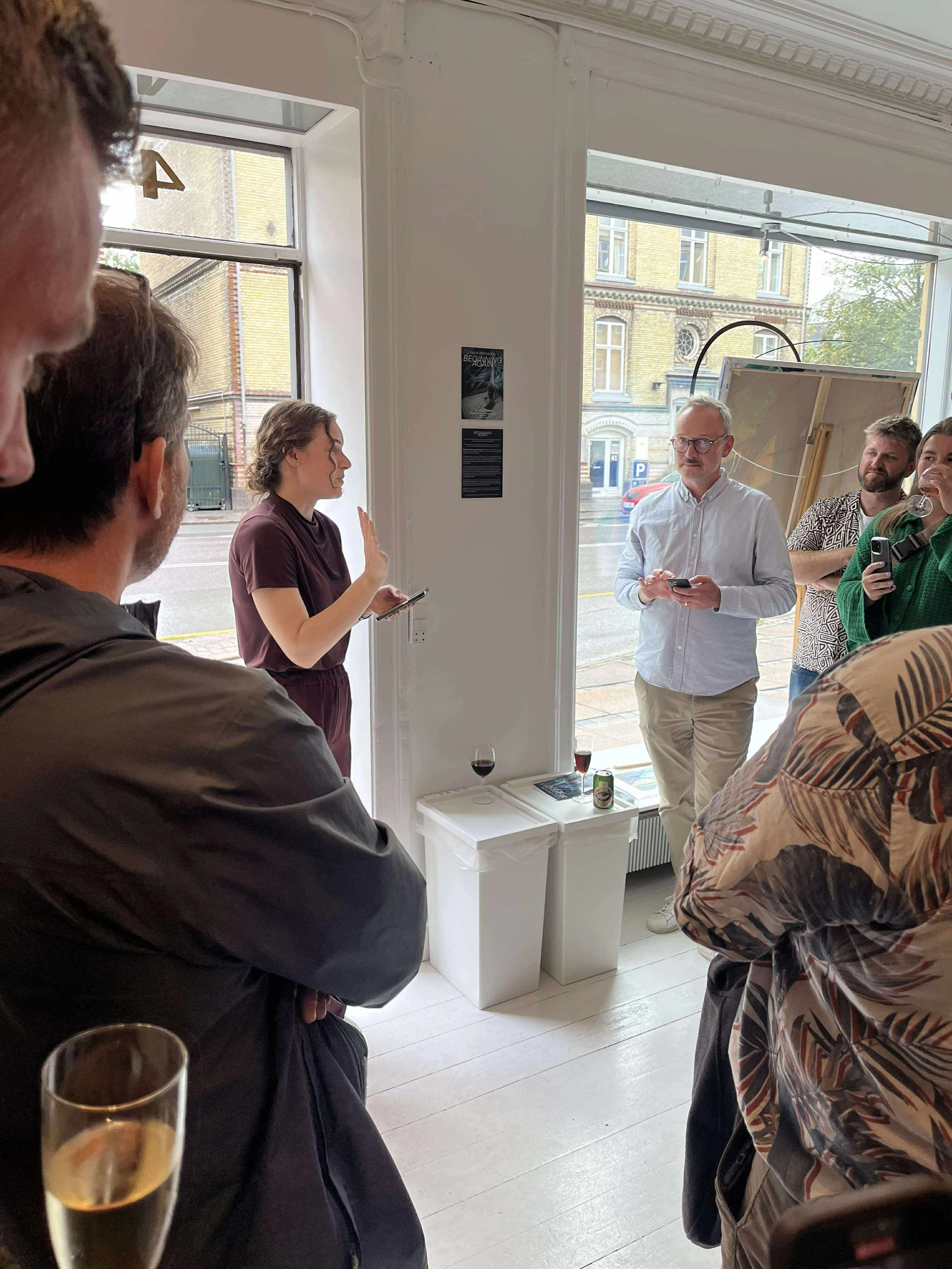 A group of people gathered inside an art gallery, some holding drinks, while Nadia Ørneborg is presenting her first excibition "Beginning Again", next to a large window showing the city streets outside.