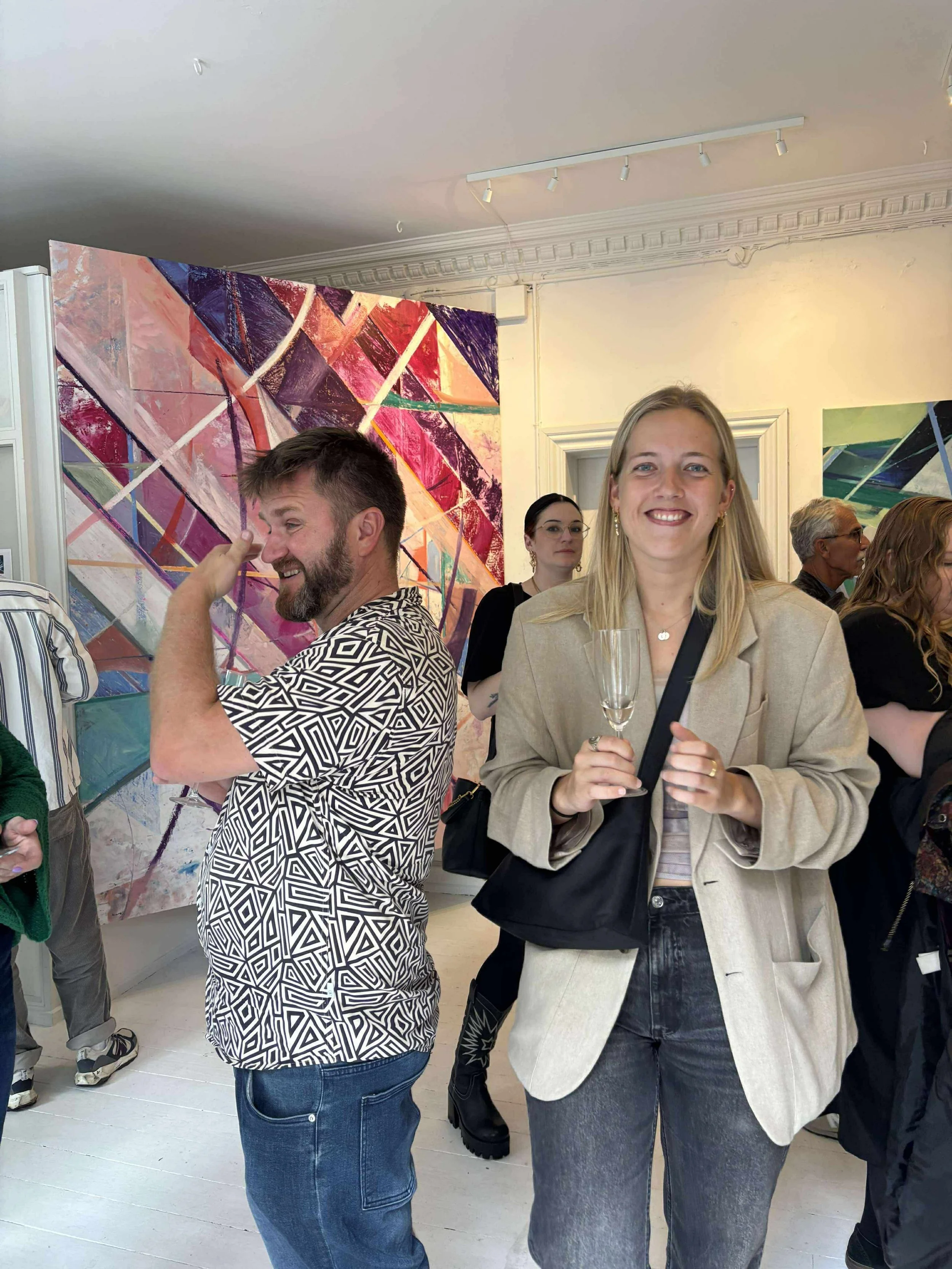 People attending an art exhibition, with a colorful abstract painting on the wall behind them. A woman in the foreground is smiling and holding a glass, wearing a beige blazer and dark jeans. A man with a beard, wearing a geometric patterned shirt, i
