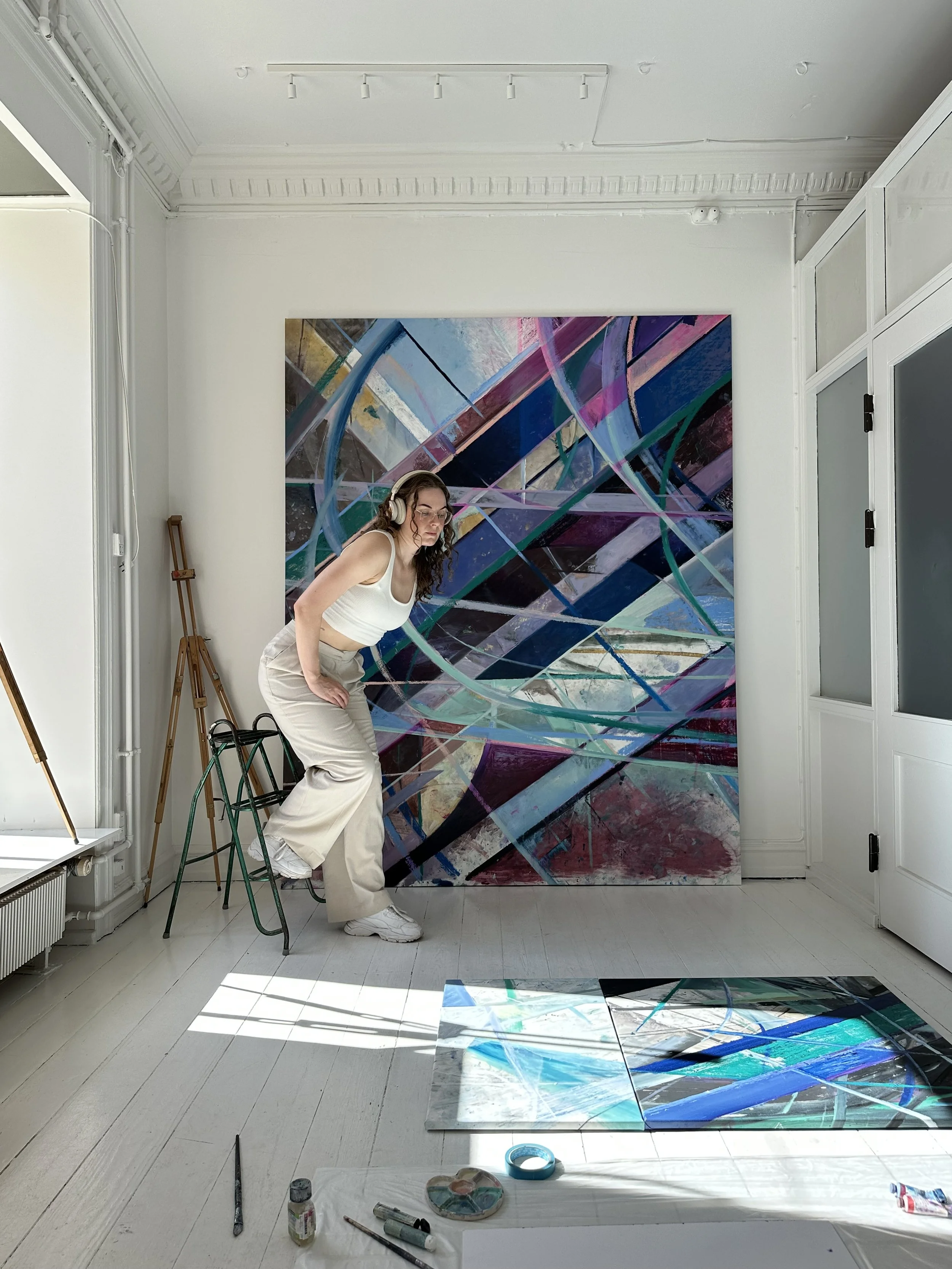 Nadia Ørneborg in a white tank top and beige pants stands up from a stool in front of her large abstract painting in a gallery.