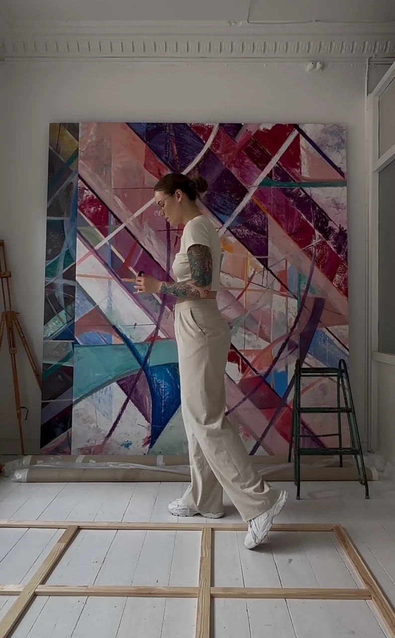 Nadia Ørneborg in full body profile wearing glasses and dressed in a white t-shirt and light-colored pants, standing in front of an abstract colourful painting, holding art materials in her hand.