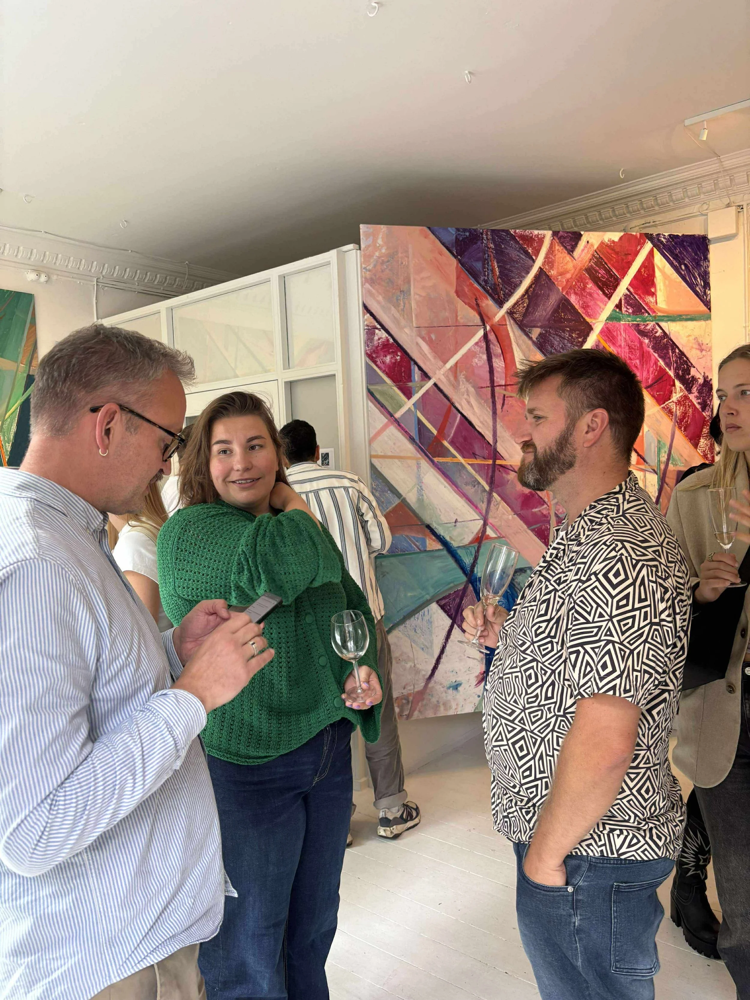 A group of people at an art gallery exhibition, mid-conversation, holding wine glasses, with a colourful abstract painting in the background.