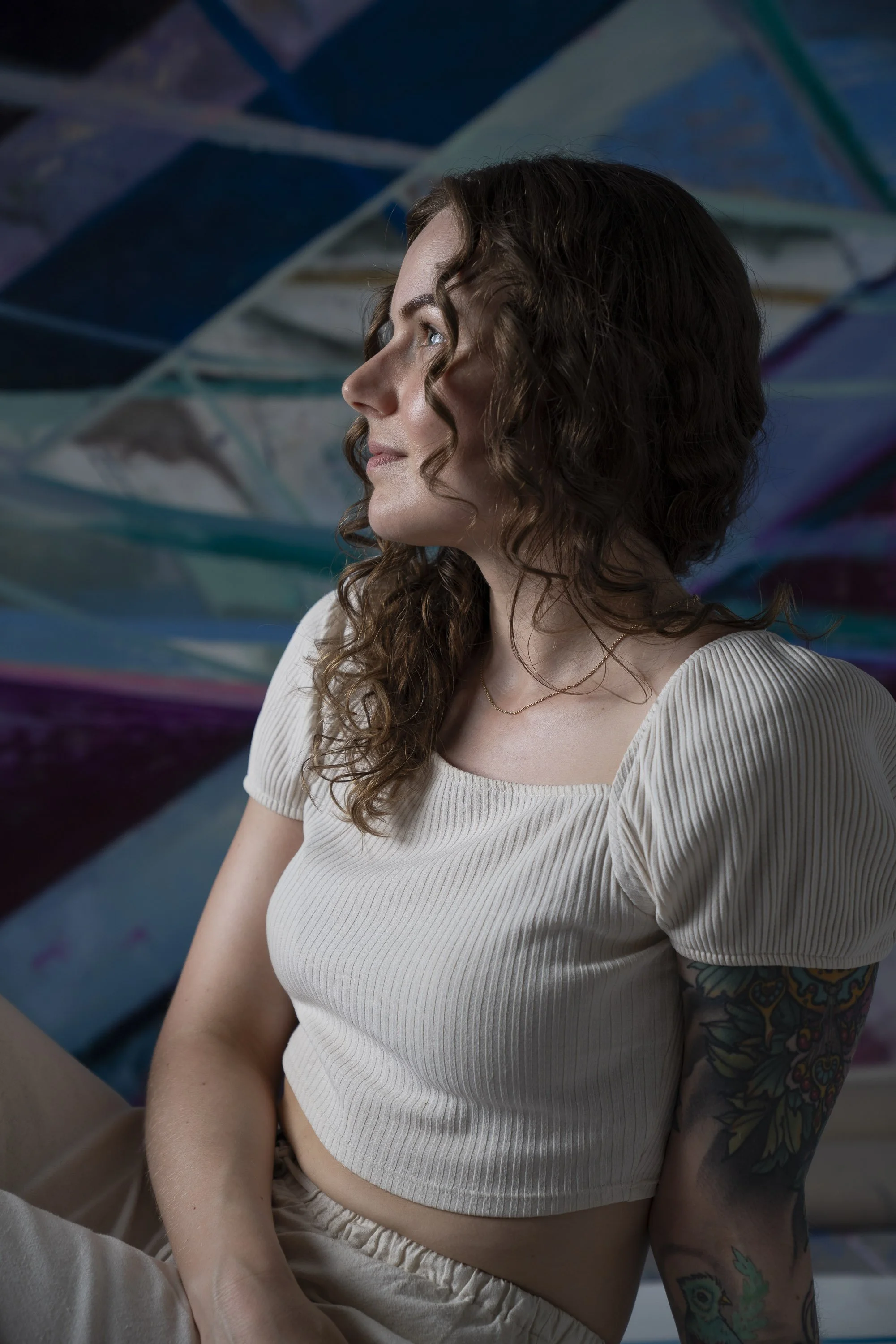 Nadia Ørneborg wearing a cream-colored ribbed crop top and beige pants, sitting on the floor looking to one side against a colorful abstract art background.