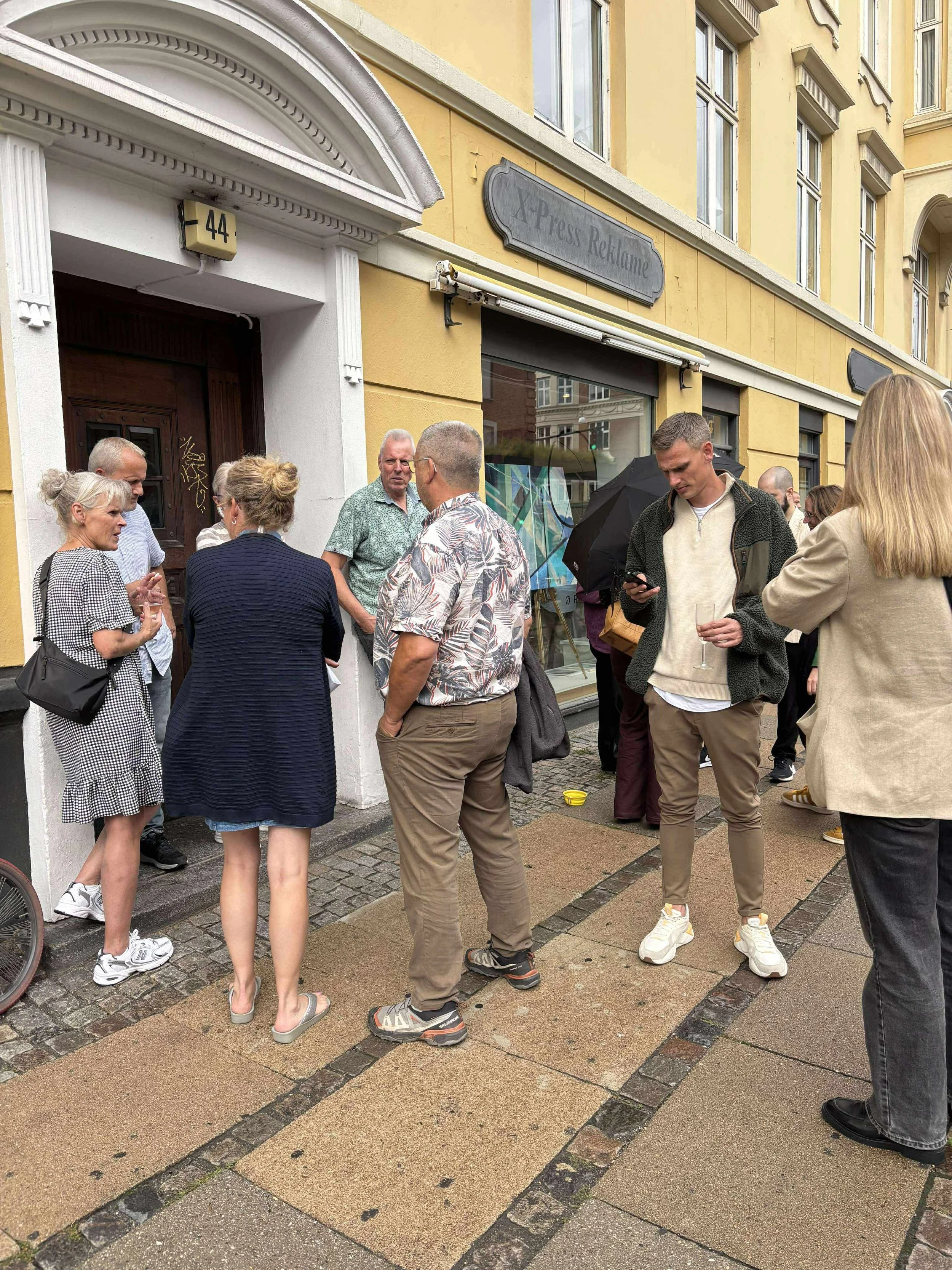 A group of people standing in front of Gallery Ørn. They are engaging in conversation and some are looking at their phones. The building is yellow with large windows.