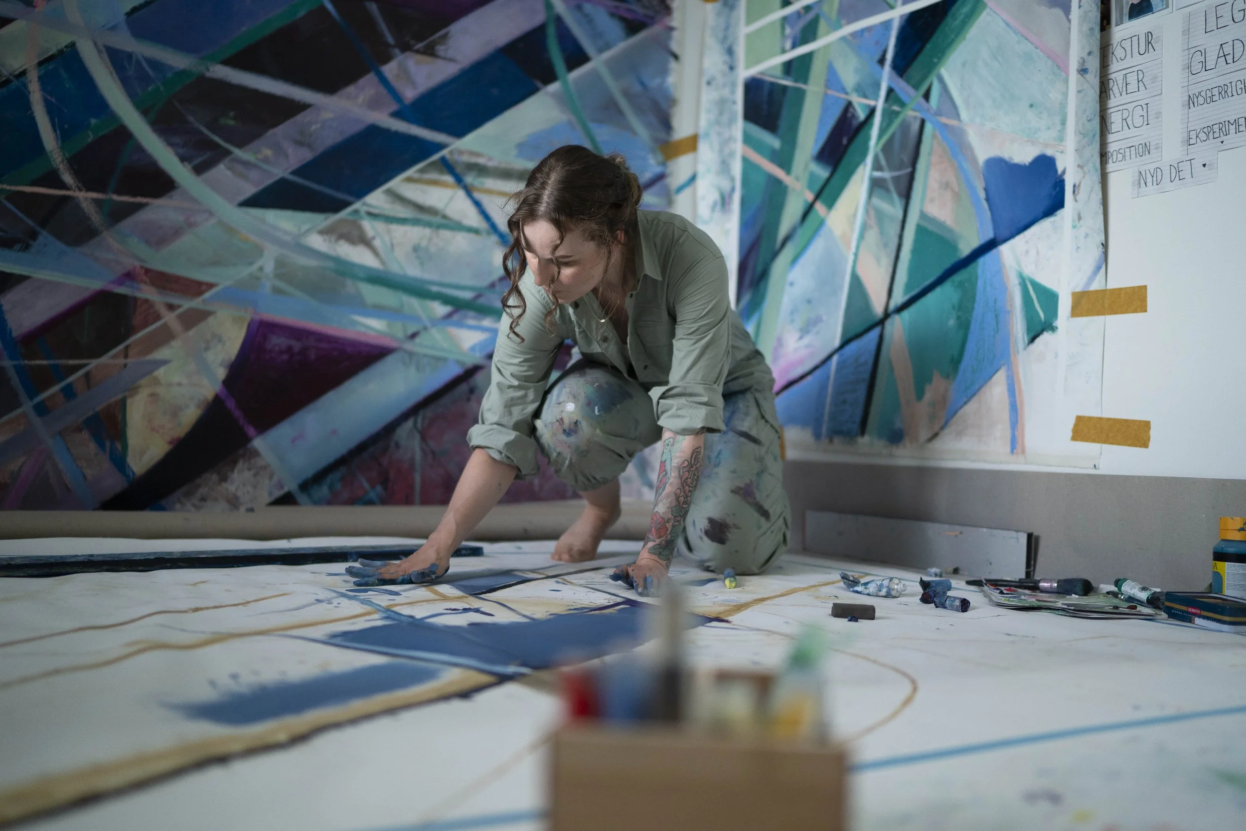 Nadia Ørneborg kneels on the floor, painting a large artwork on a white canvas. She is painting n her living room with abstract colourful artwork hanging on the wall behind her.