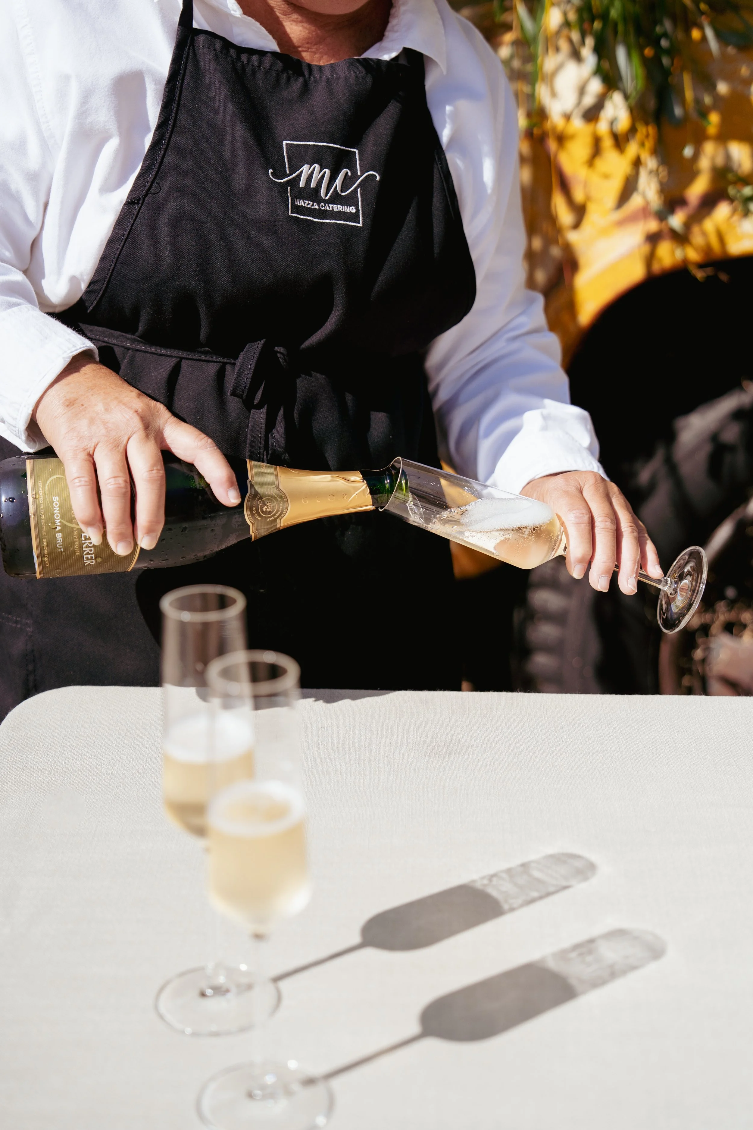 A person pouring champagne into a flute glass at an outdoor event, with two more filled champagne flutes on a table in front. The person is wearing a black apron with a logo and a white shirt.