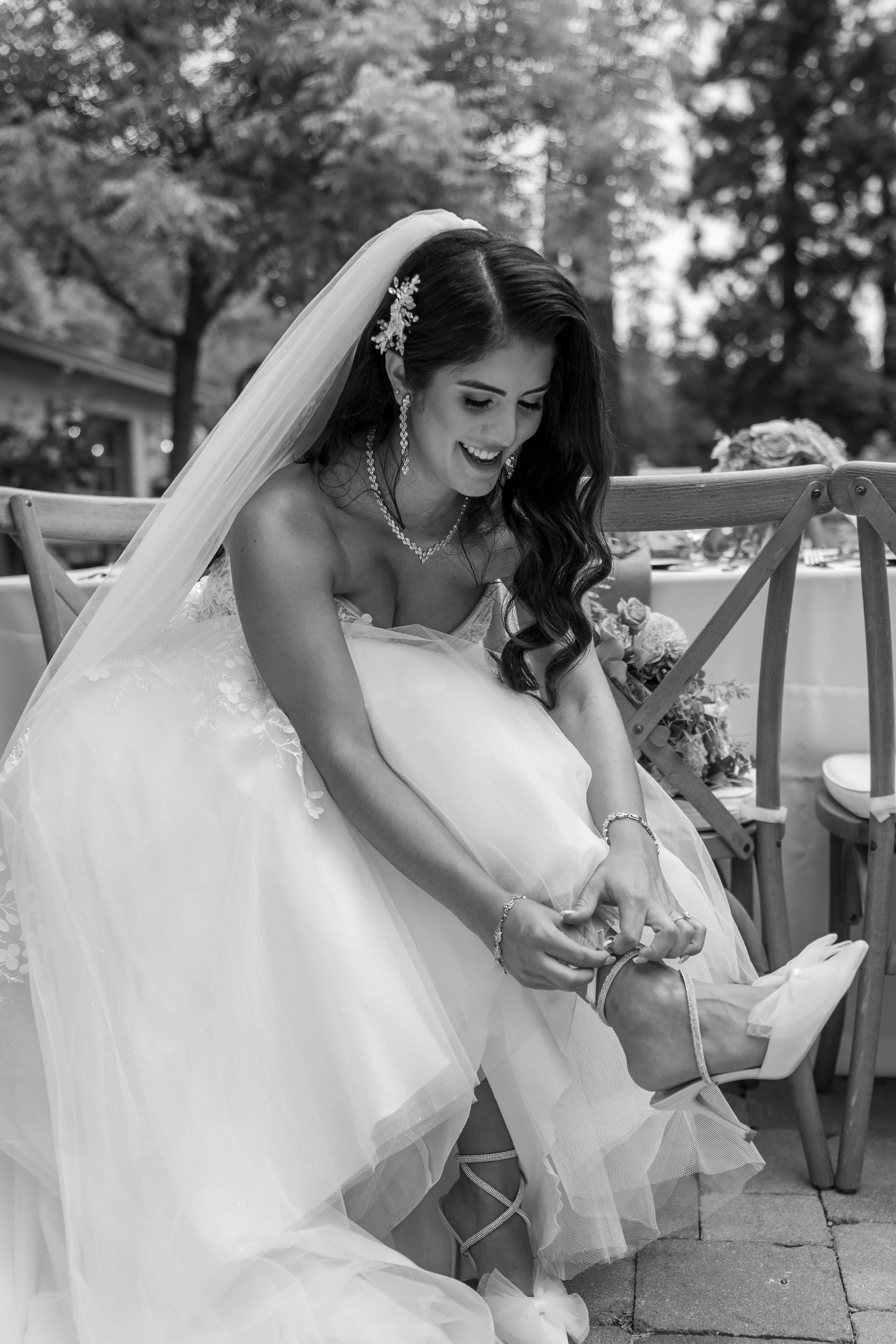 A woman in a wedding dress with a veil is smiling and sitting while putting on her high-heeled shoes at a wedding reception outdoors, with trees and tables in the background.