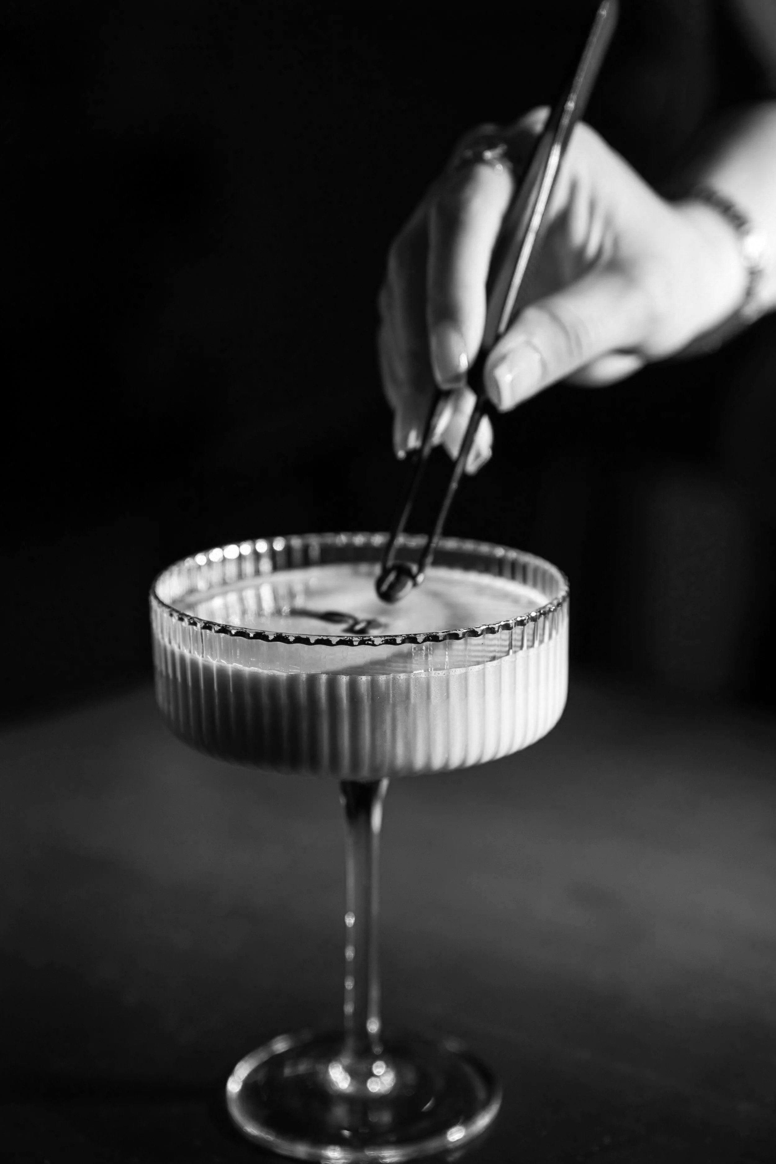 Black and white photo of a hand holding tongs, reaching into a glass coupe with a layered drink, on a dark surface.