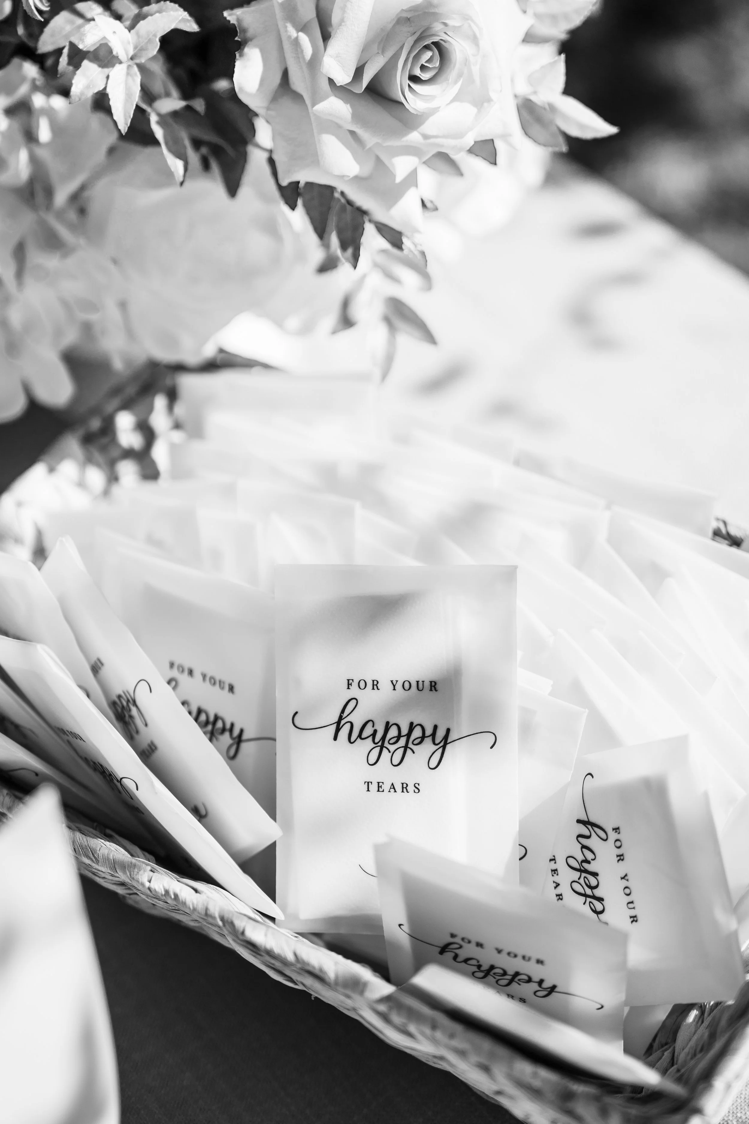 Black and white photo of small gift bags with the text "For your happy tears" printed on them, arranged in a basket beneath a bouquet of roses and other flowers.