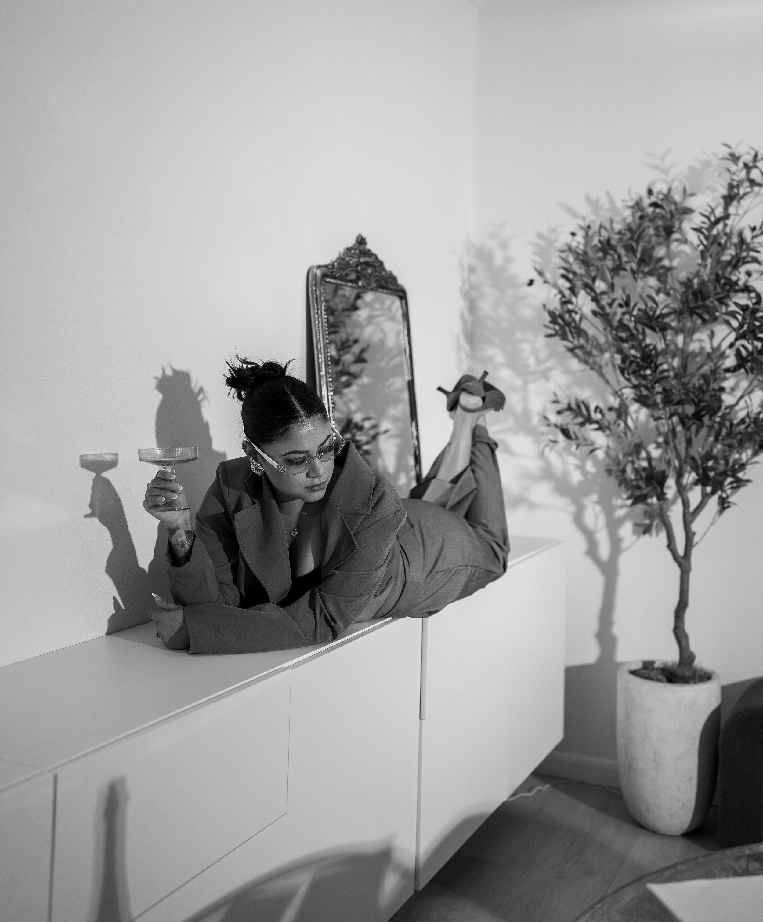 Black and white photo of a woman in professional attire lying on a white cabinet, holding two glasses and looking down, with a mirror, a potted tree, and shadows on the white wall in the background.