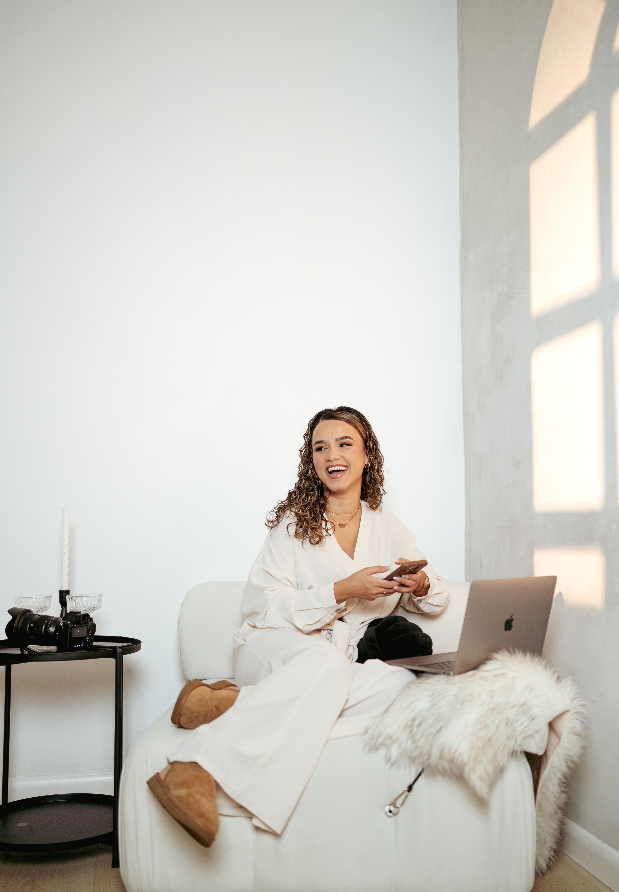 A woman with curly hair, wearing a white outfit, sitting on a beige sofa, smiling and looking at a smartphone, with a laptop in front of her, in a bright room with sunlight casting shadows through a window.