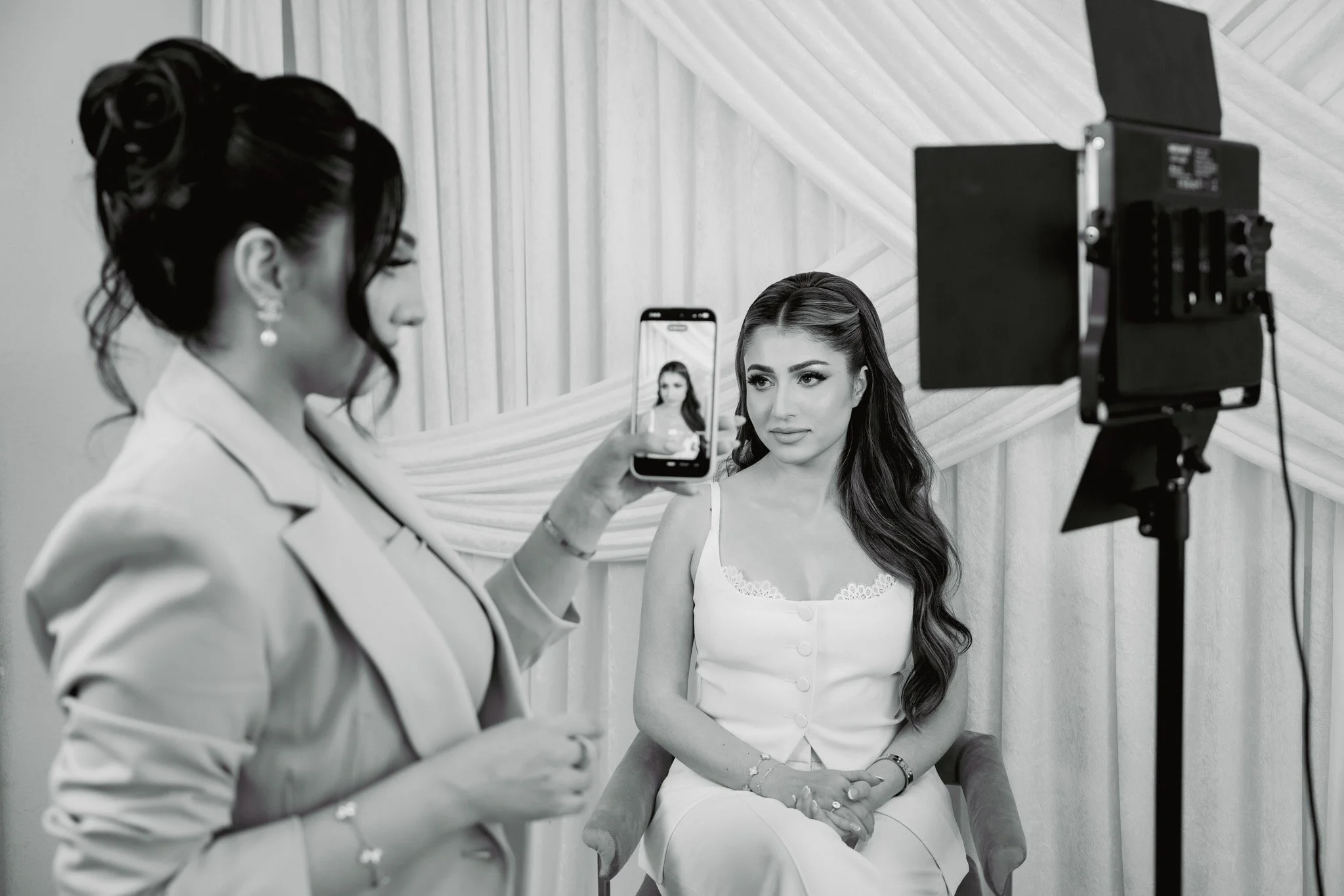 A woman is sitting on a chair while another woman takes her photo with a smartphone during a photo shoot in a studio with draped curtains in the background.