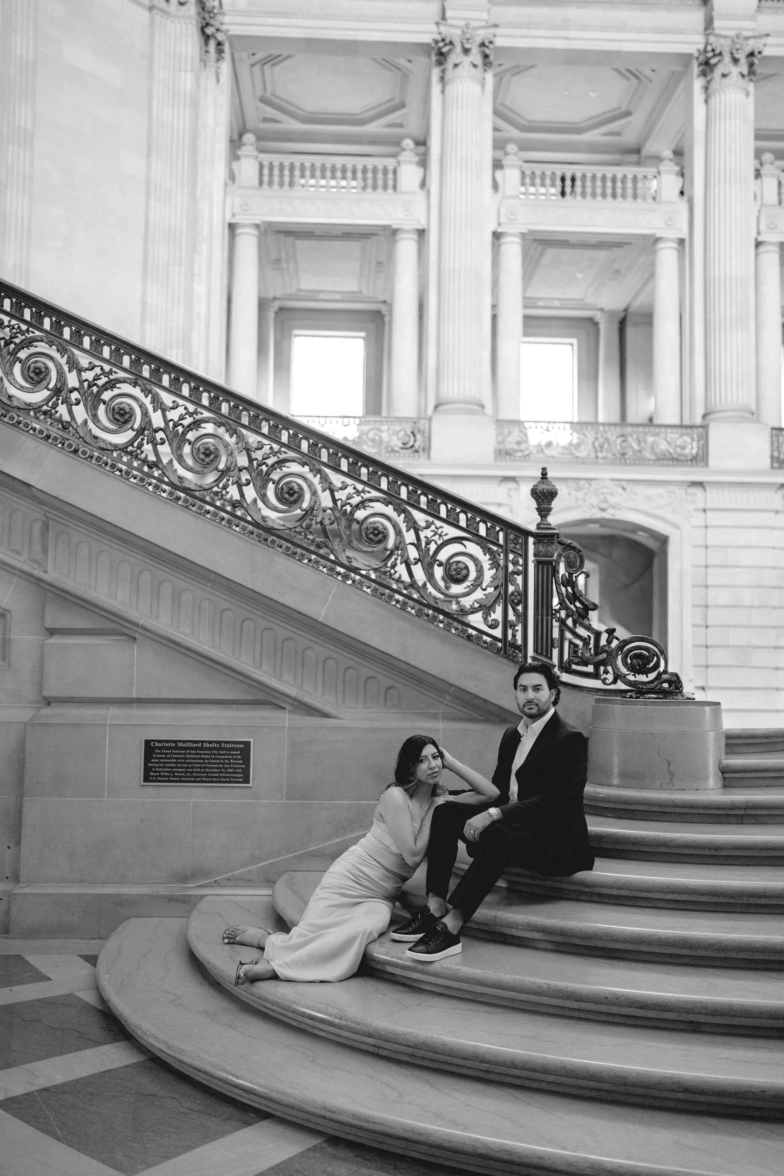 A man in a suit and a woman in a dress sitting on the steps of a grand, ornate staircase in a historic building.