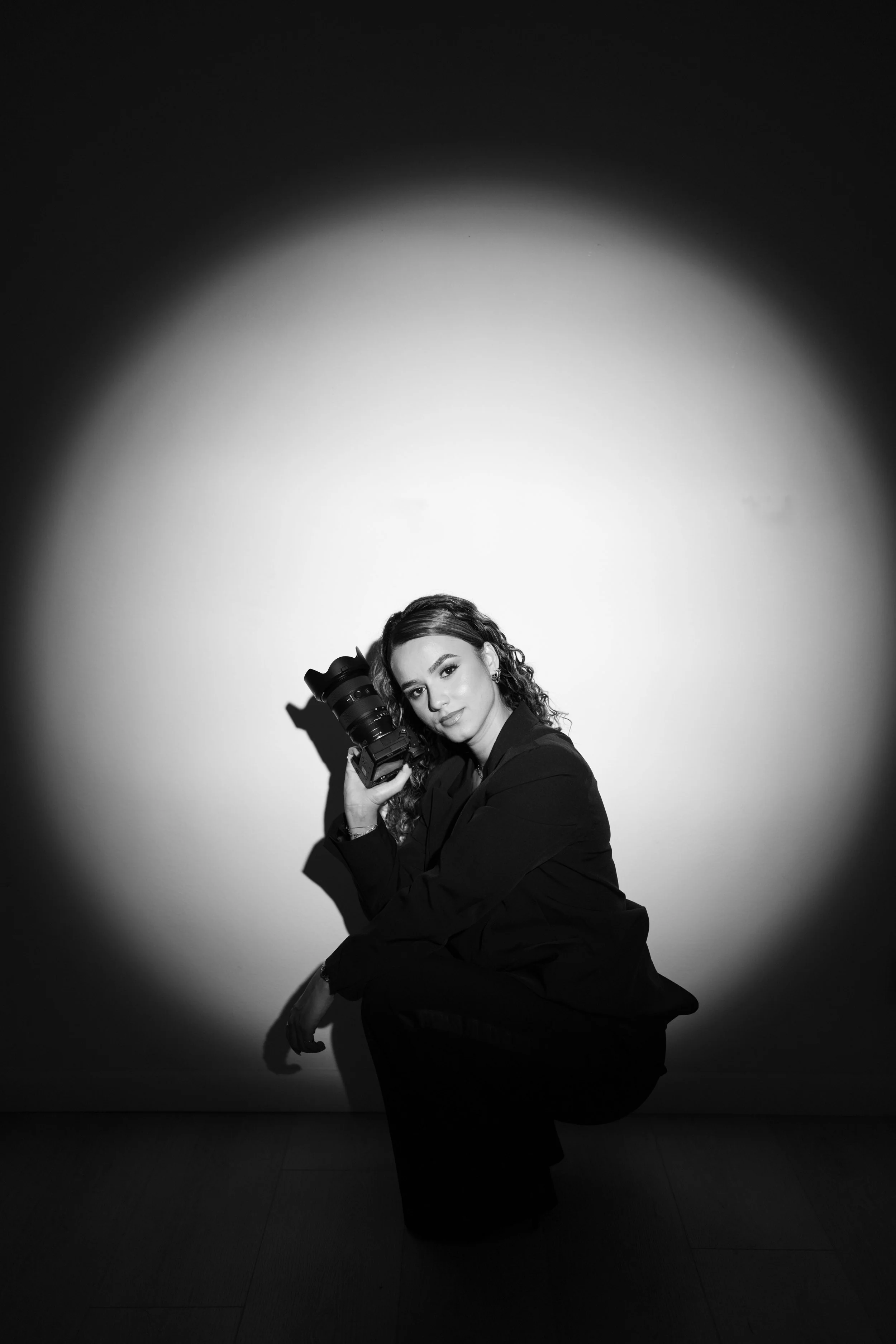 Black and white photo of a woman crouching, holding a professional camera, with a spotlight illumination creating a circular light pattern on the wall behind her.