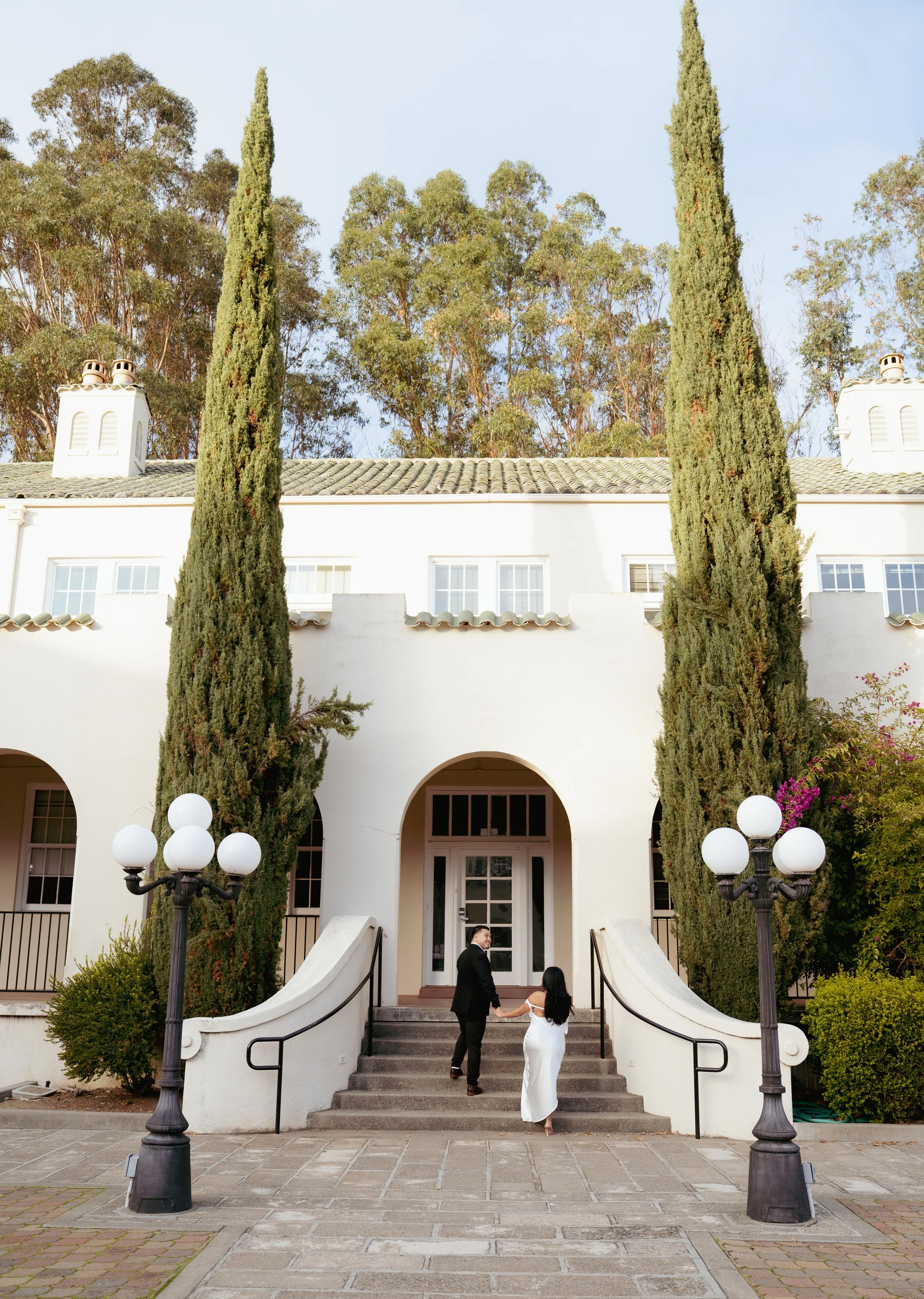 A man and woman holding hands on the steps of a white building with arched entrance, tall cypress trees, lamp posts, and greenery.