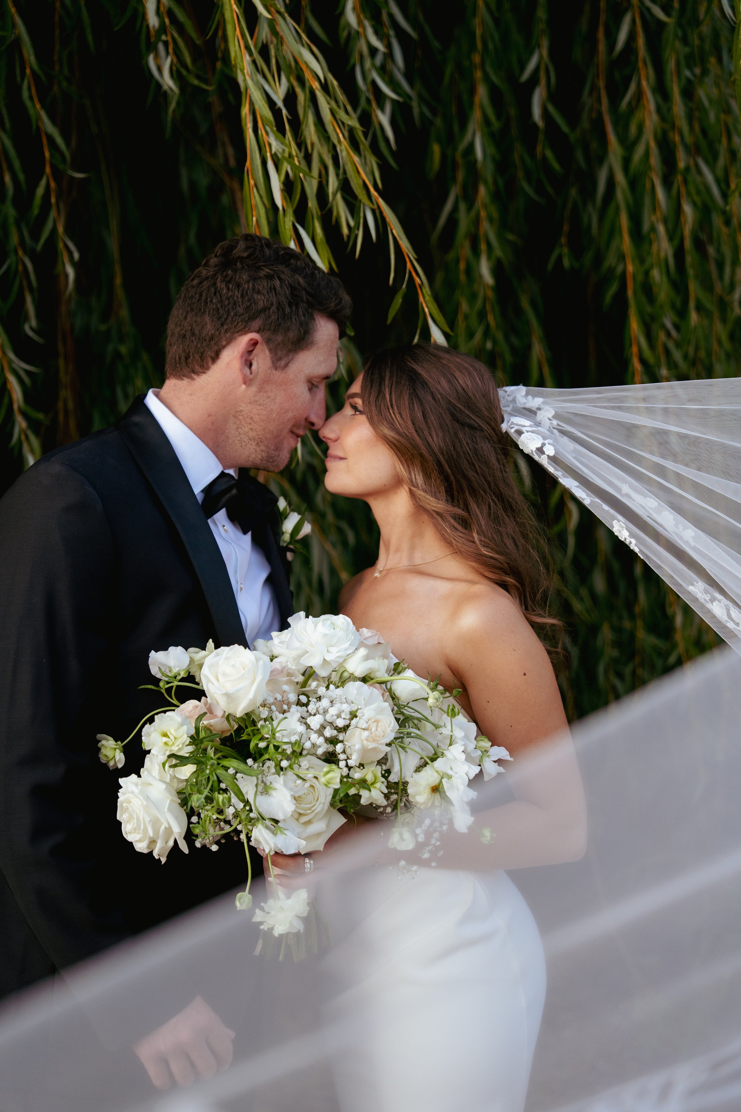 A bride and groom standing close together, touching foreheads and smiling, with a lush green background. The bride holds a large bouquet of white flowers, and is wearing a strapless wedding dress with a veil. The groom is dressed in a black tuxedo with a white shirt and black bow tie.