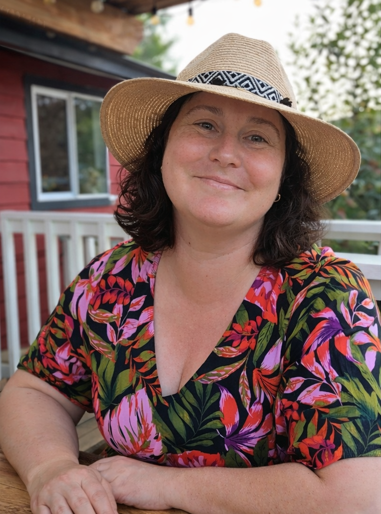 A woman smiling, wearing a colorful floral dress and a wide-brimmed straw hat, sitting outdoors on a deck with a white railing and a red building with windows in the background.