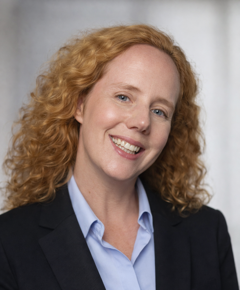 A woman with curly red hair wearing a black blazer and light blue collared shirt, smiling at the camera.