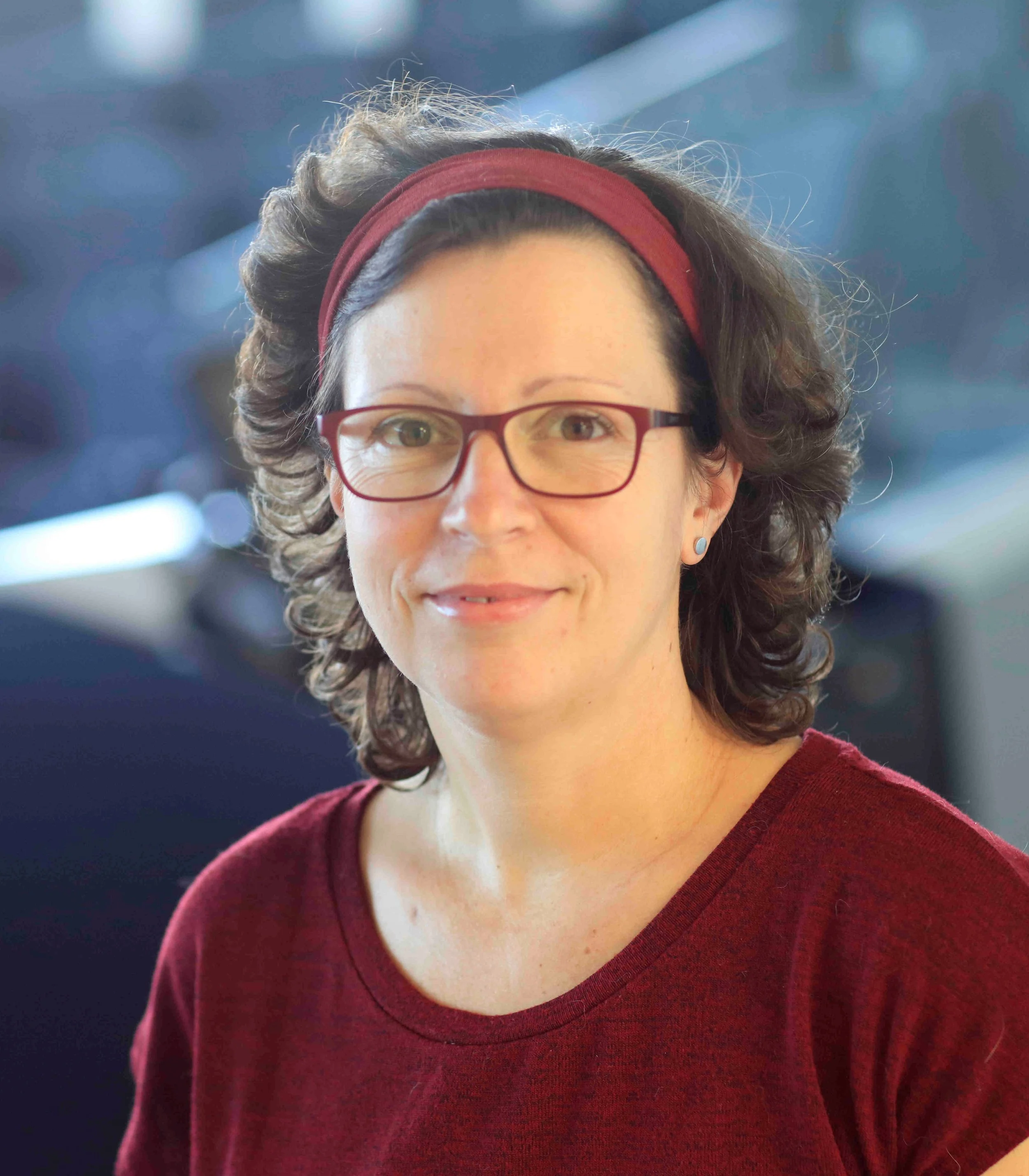 A woman with curly brown hair, wearing glasses and a red headband, smiling in a close-up portrait.