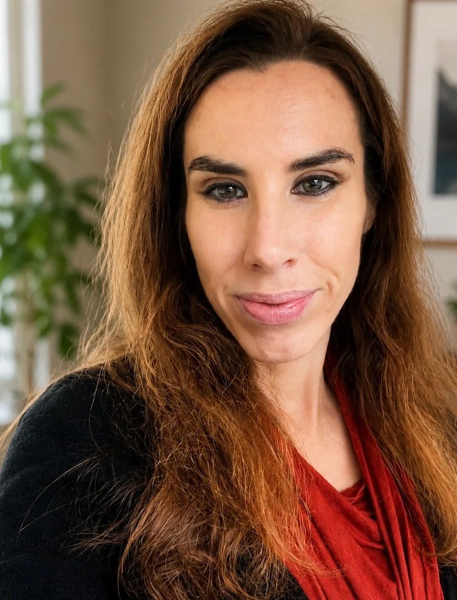 Close-up of a woman with long, wavy brown hair, wearing a black top and a red scarf inside a room with a blurred background of a plant and framed pictures.