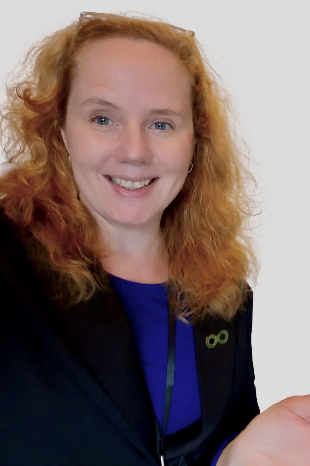 A woman with curly red hair and wearing glasses on her head, smiling at the camera, dressed in a black blazer and a blue top, against a plain white background.