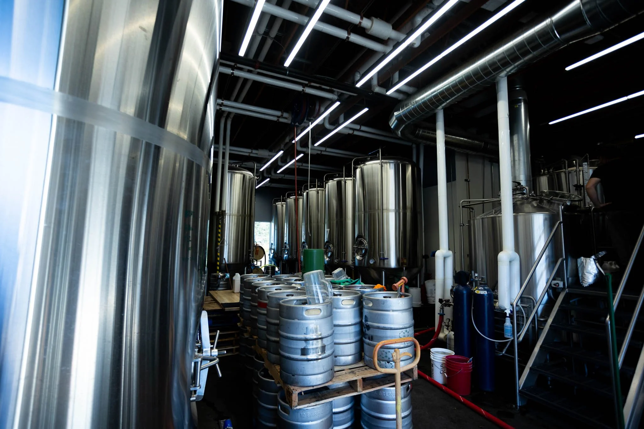 Inside a brewery with large stainless steel fermentation tanks and stacked beer kegs on a pallet.