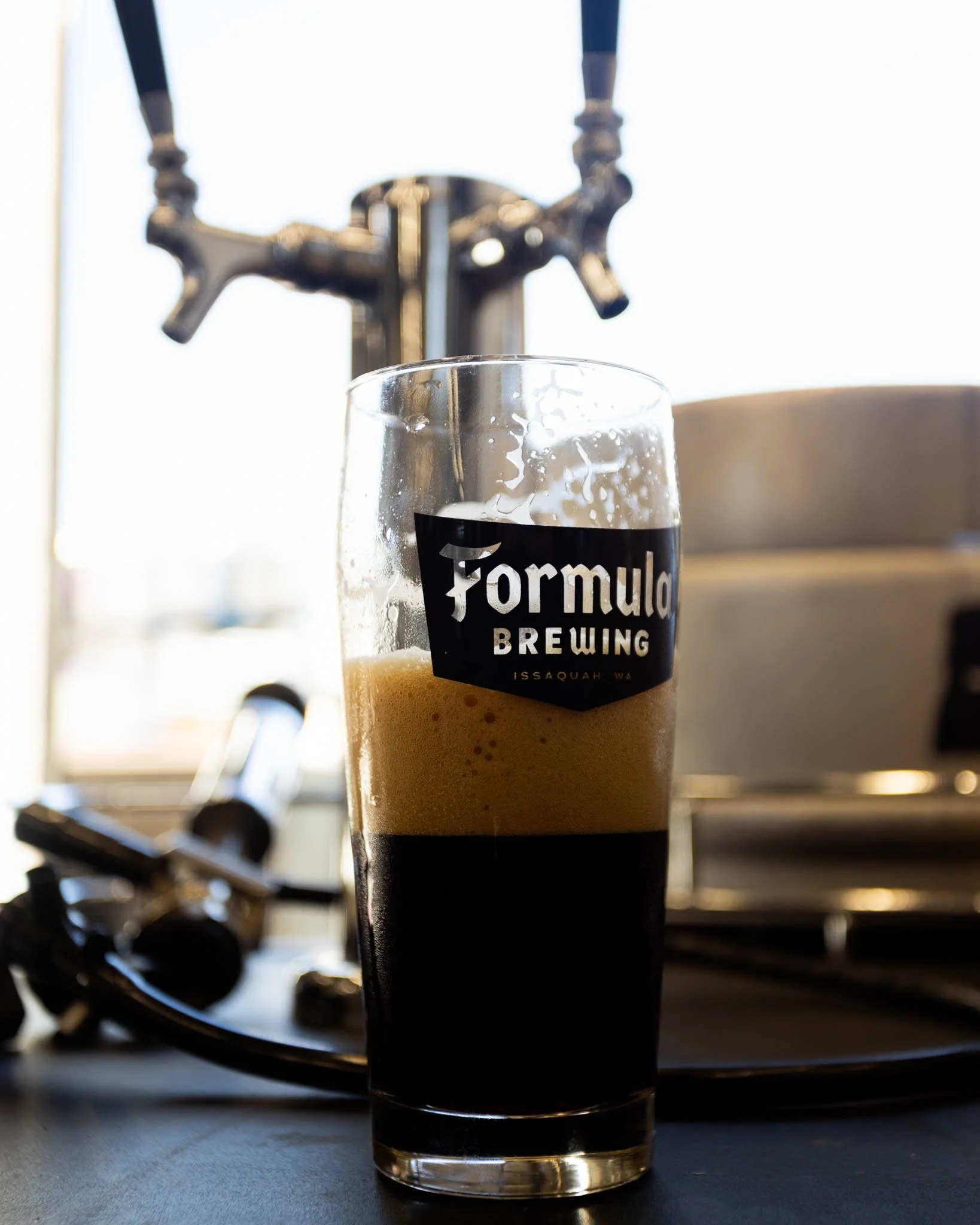 A glass of dark beer with foam on top, labeled 'Formula Brewing' from Issaquah, Washington, sits on a table in front of a brewing tap system.