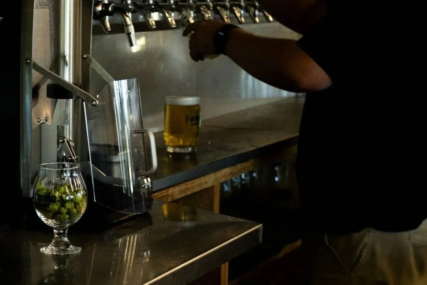 Person pouring beer from tap into a glass at a bar, with a glass of beer on the counter and a jar of green olives nearby.