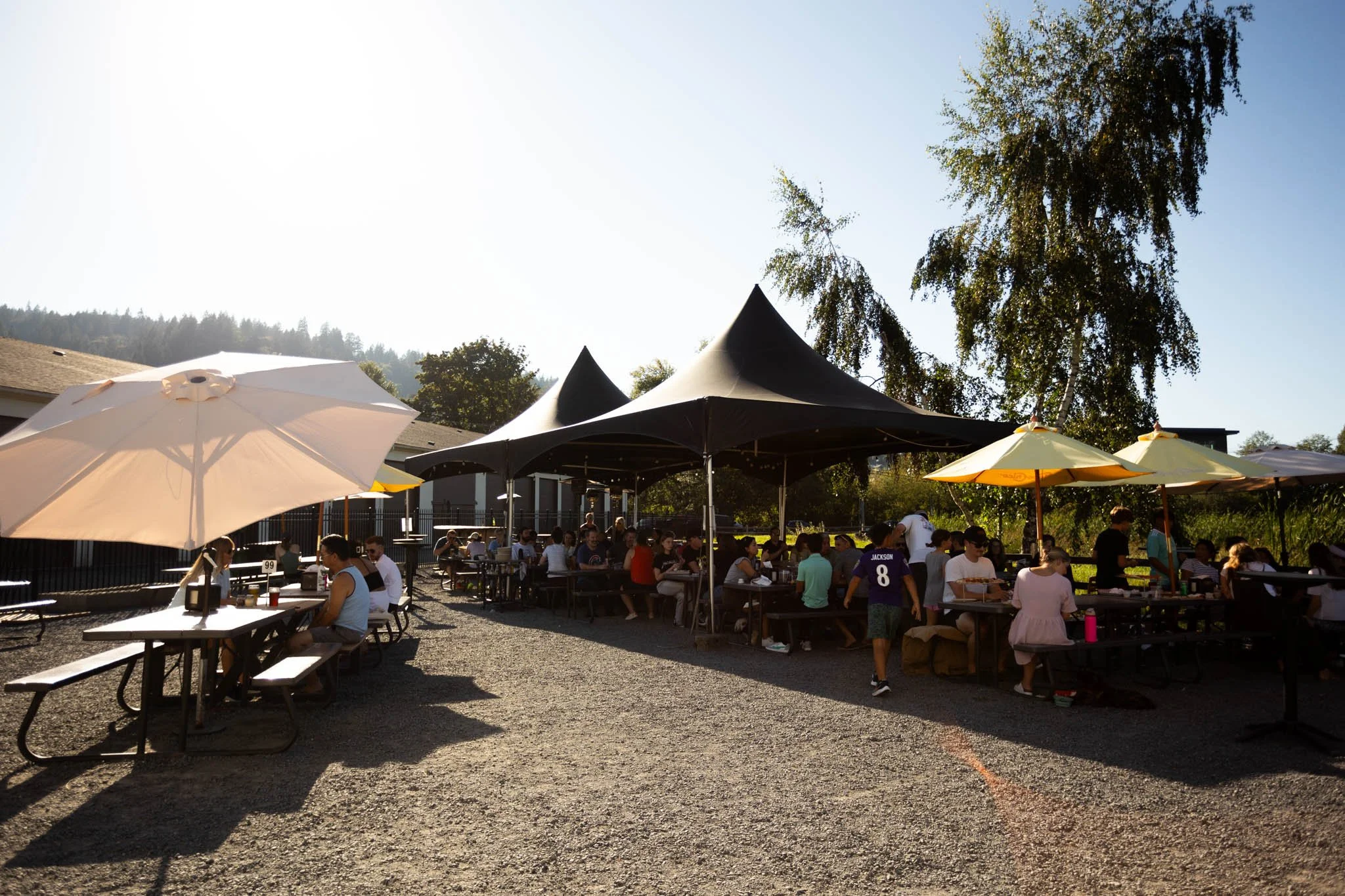 People sitting and socializing at outdoor tables with umbrellas, under tents, in a gravel area near trees and a building, during daytime.