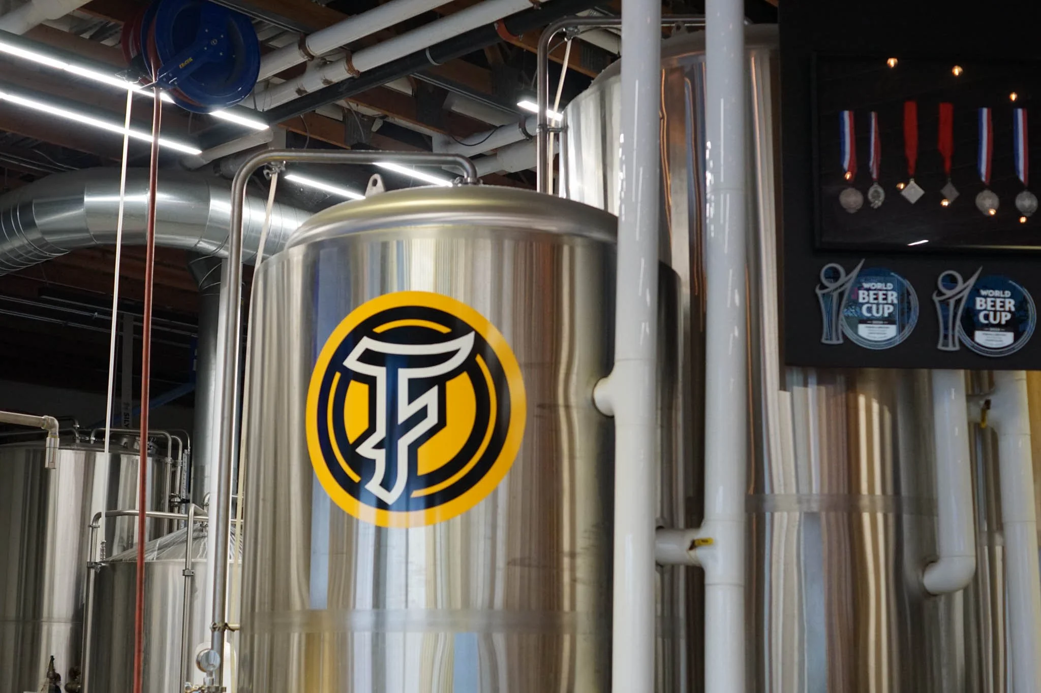 Large stainless steel beer fermentation tank in a brewery, with a logo featuring a stylized pint glass and the letter T in black and white on a yellow circle.