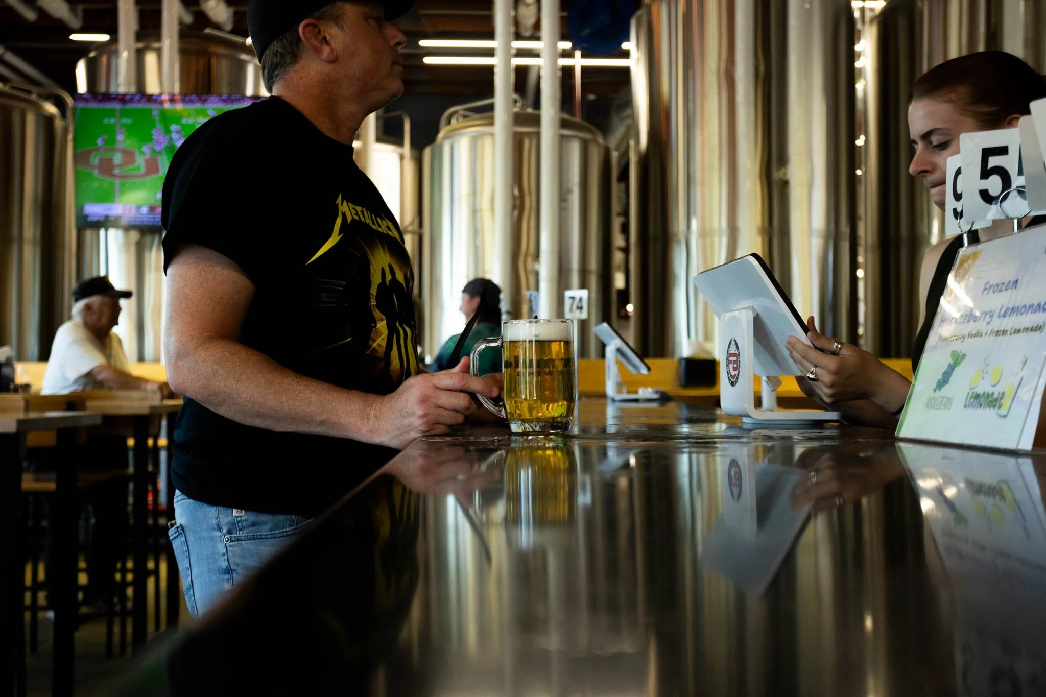 A man with a Metallica T-shirt at a bar counter holding a glass of beer, talking to a female bartender using a tablet, inside a brewery with stainless steel tanks.