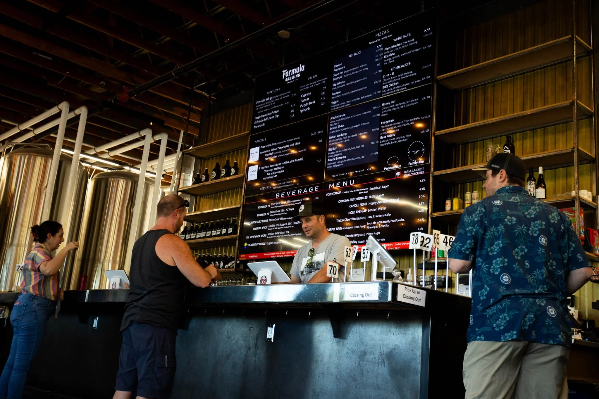 People ordering drinks at a brewery counter with a large digital menu board displaying pizza options and beverages.