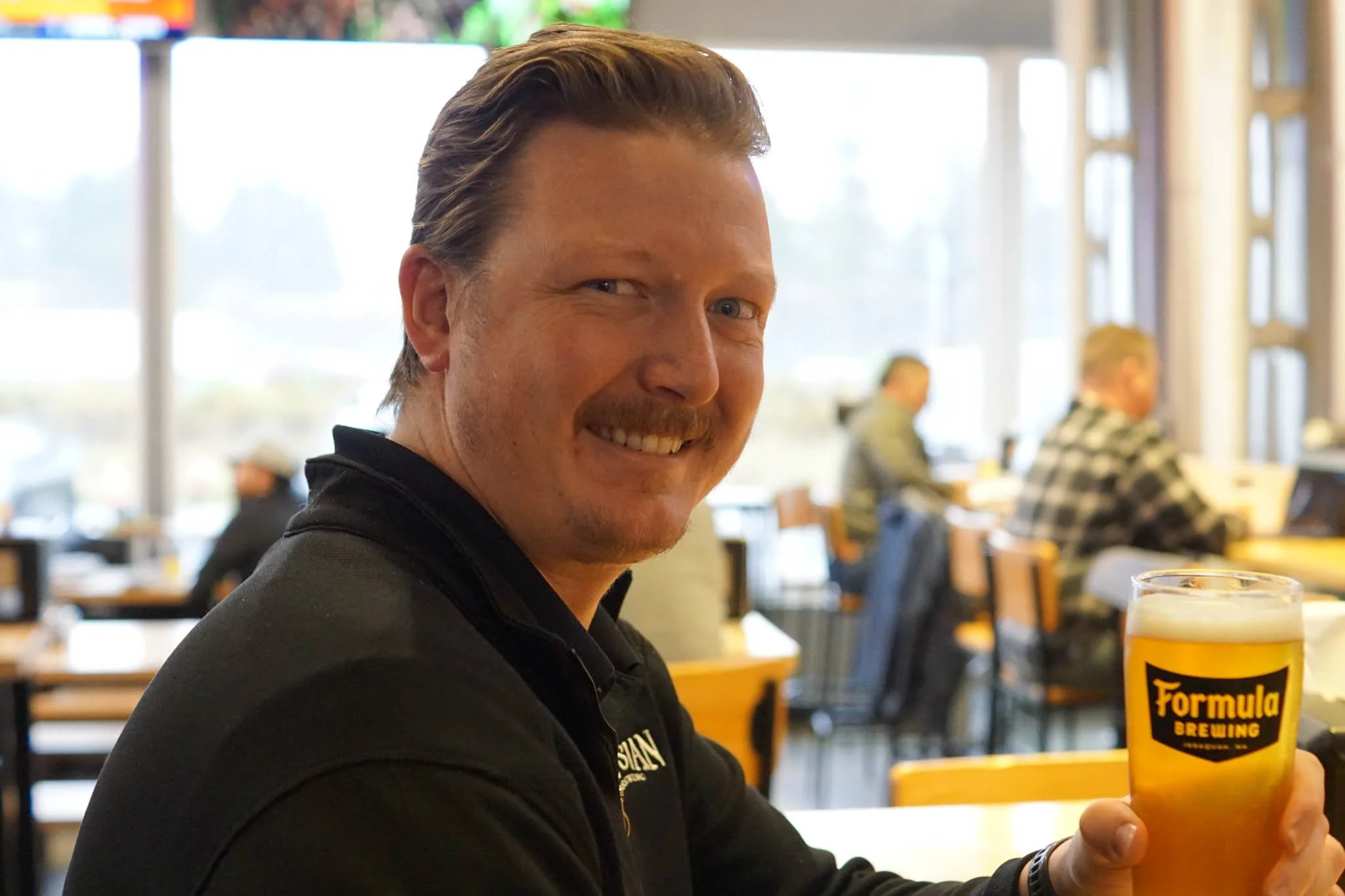 A man smiling and holding a glass of beer in a brewery or pub with large windows and other people in the background.