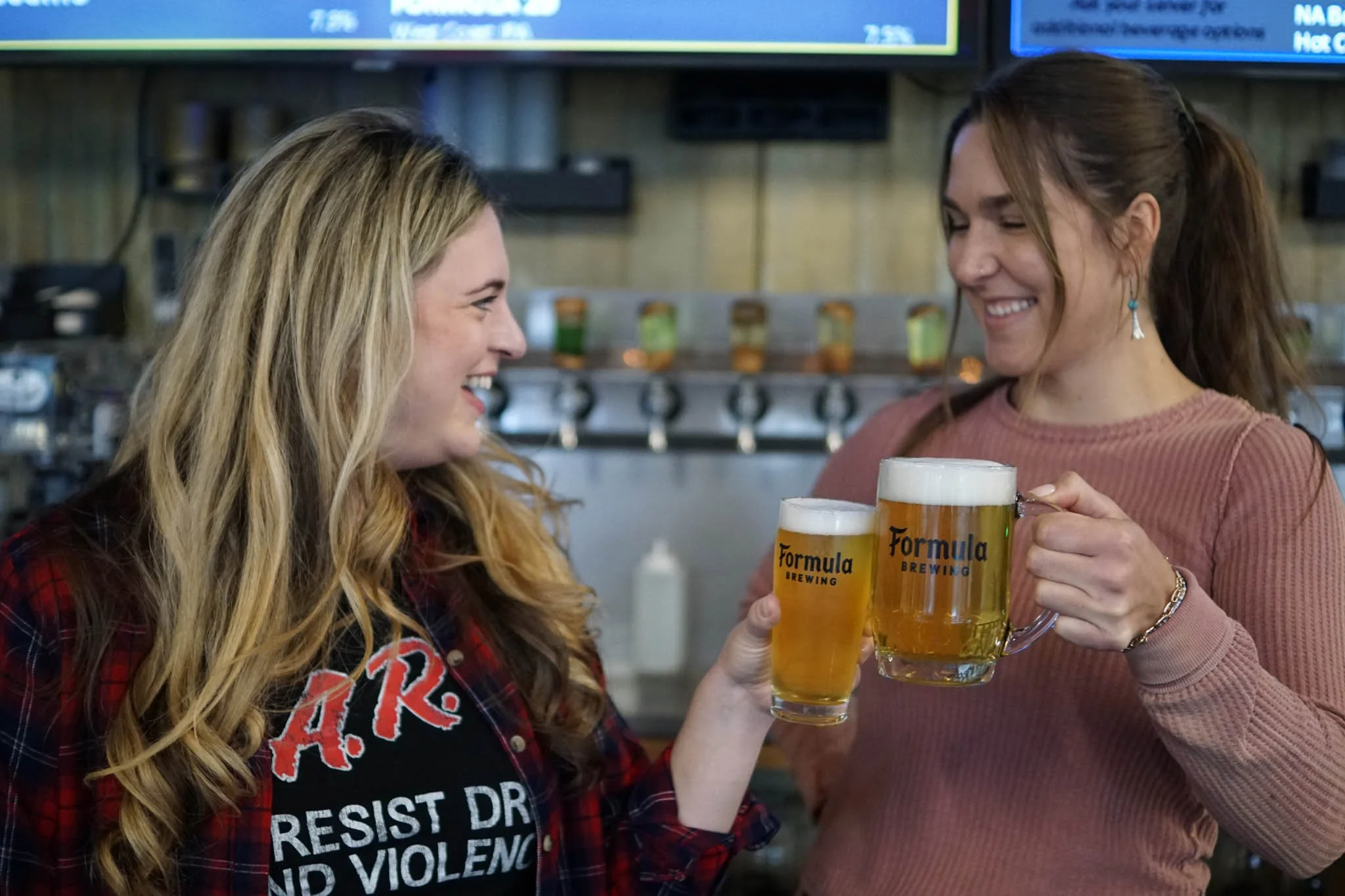 Two women smiling and clinking glasses of beer at a bar, with the logo of Formula Brewing on the glasses.