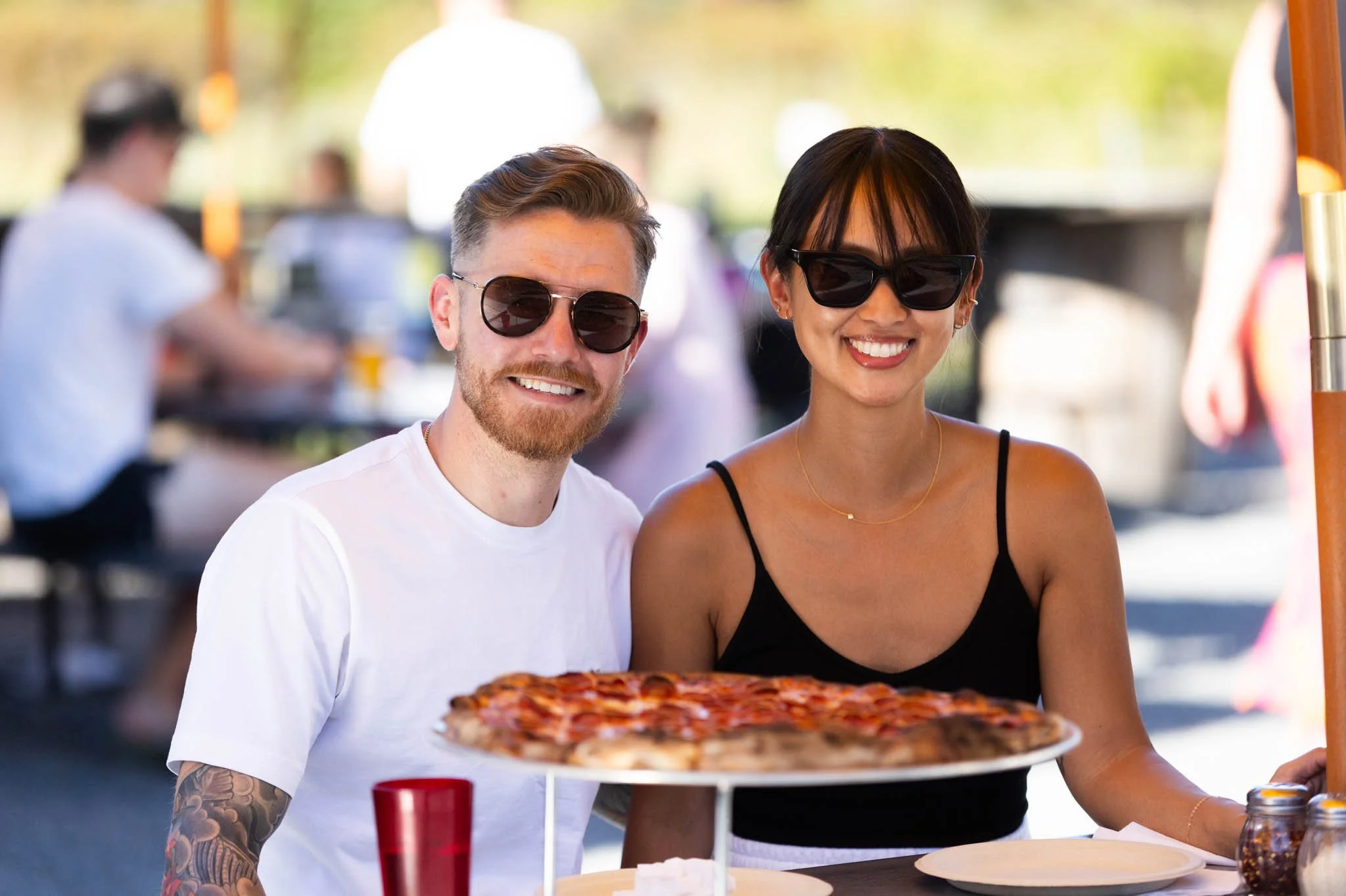 A smiling man and woman wearing sunglasses are sitting at an outdoor table, with a large pizza in front of them.