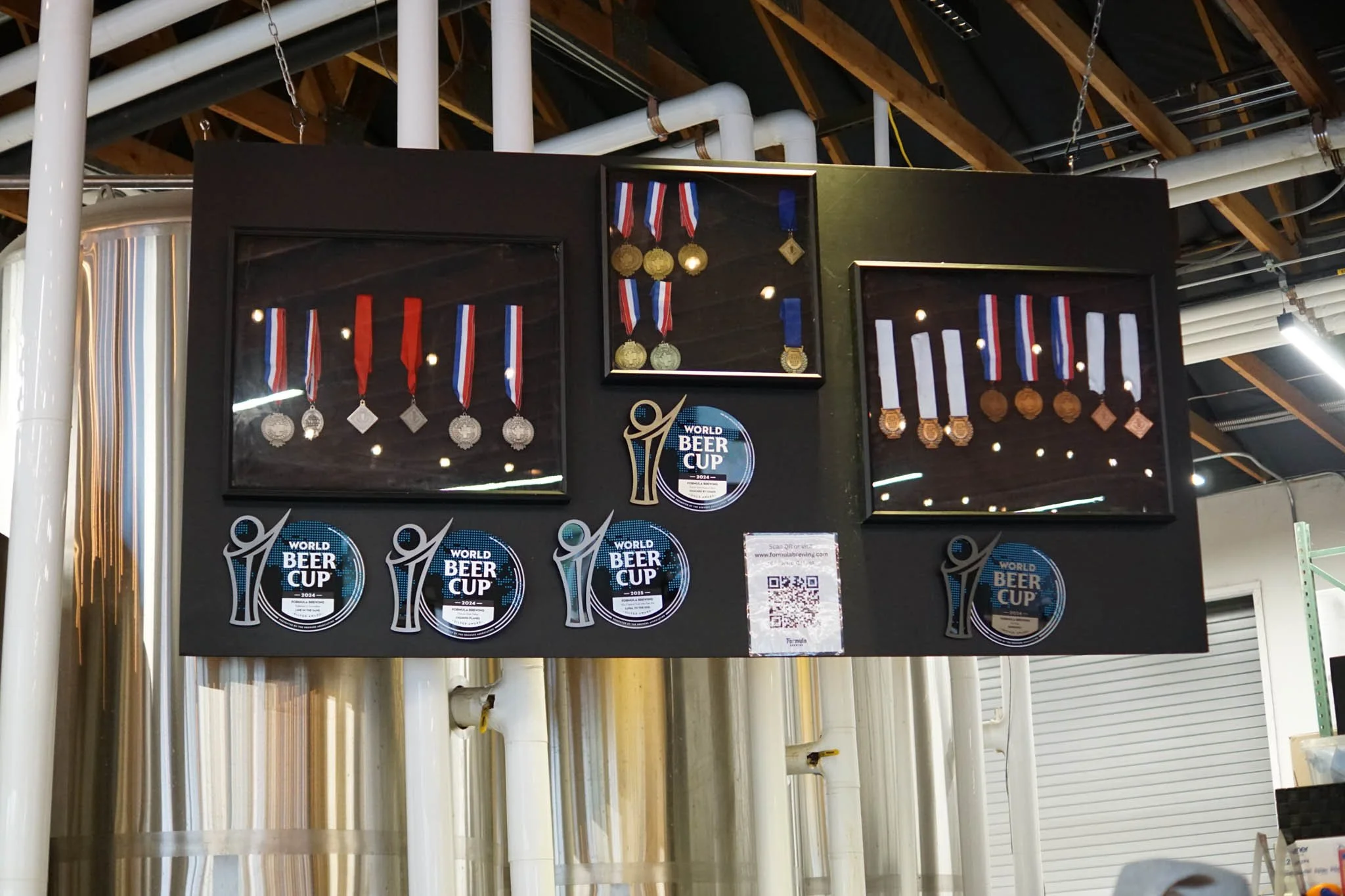 Display case with various medals, trophies, and stickers related to the World Beer Cup, set against a background of industrial pipes and wooden roof beams.