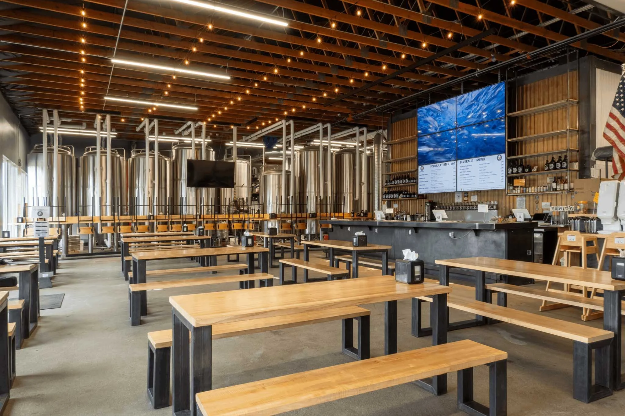 Empty brewery taproom with wooden tables and benches, large stainless steel fermentation tanks, digital menus on screens, and an American flag.
