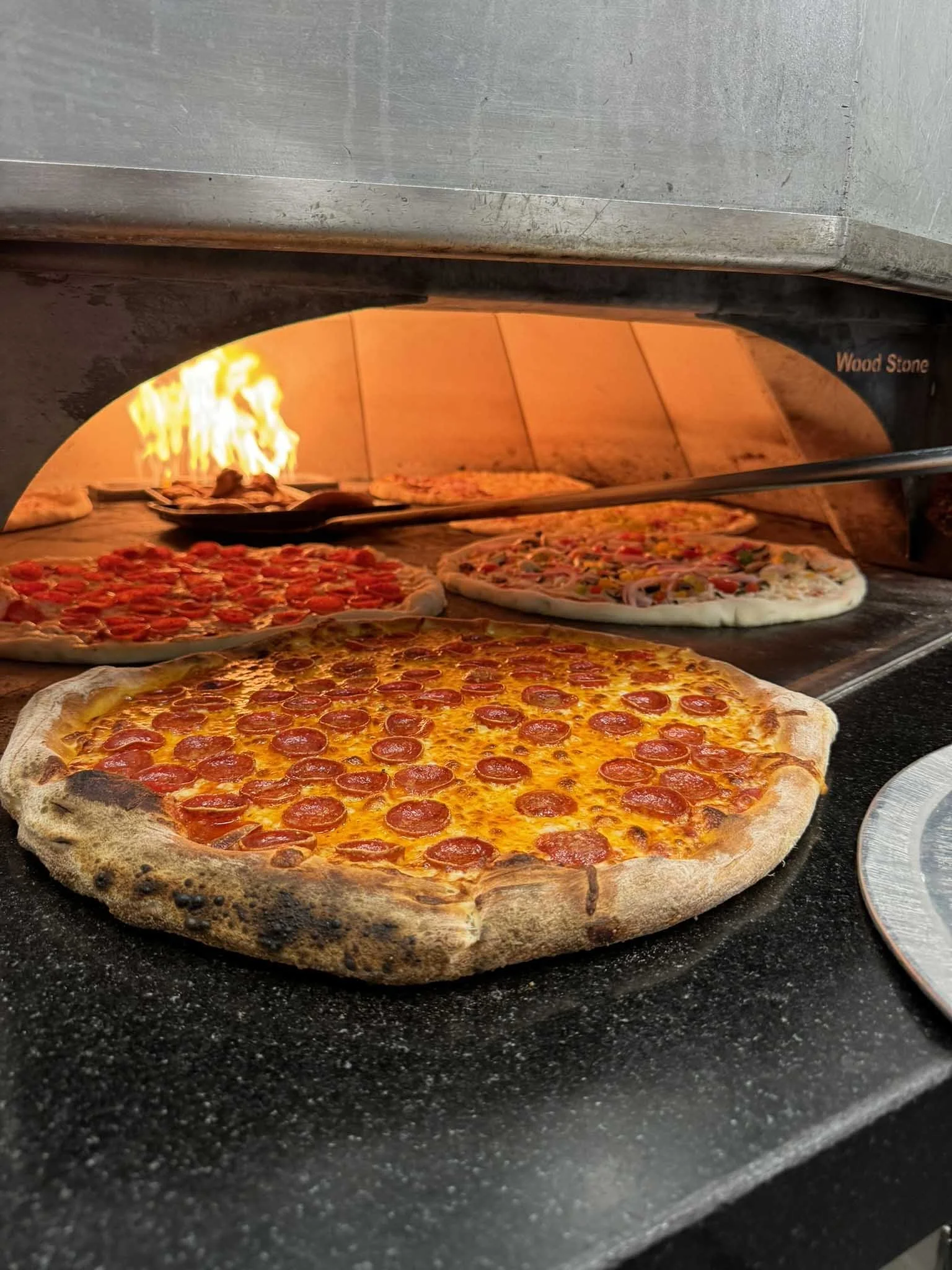 Pepperoni pizza baking in a wood-fired oven with multiple pizzas inside.