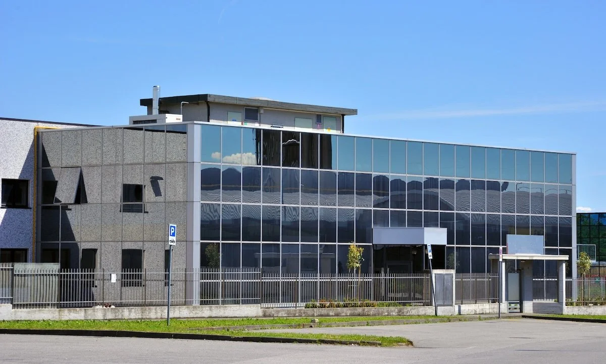 Modern building with gray concrete and black glass exterior, surrounded by a fence and parking area.
