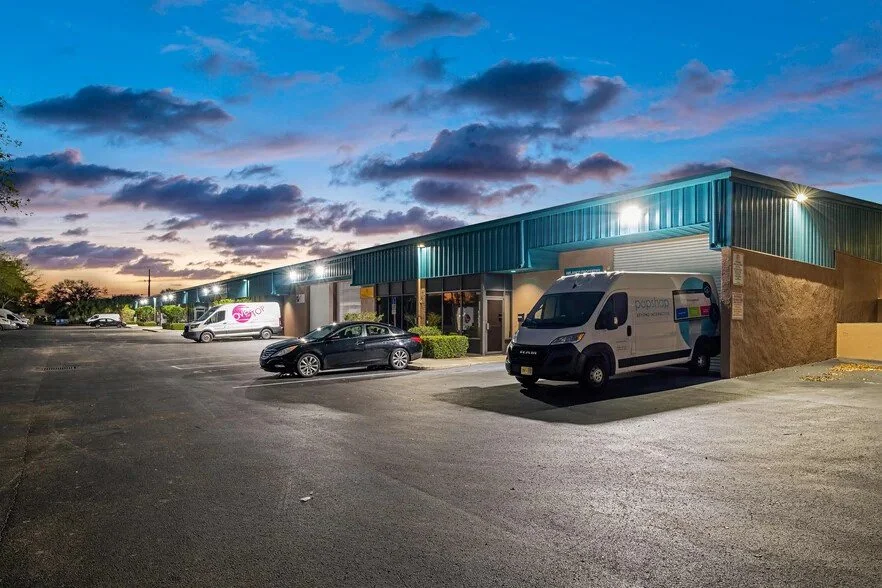Photo of a commercial building with a parking lot at dusk, featuring several parked cars and vans, with a partly cloudy sky overhead.