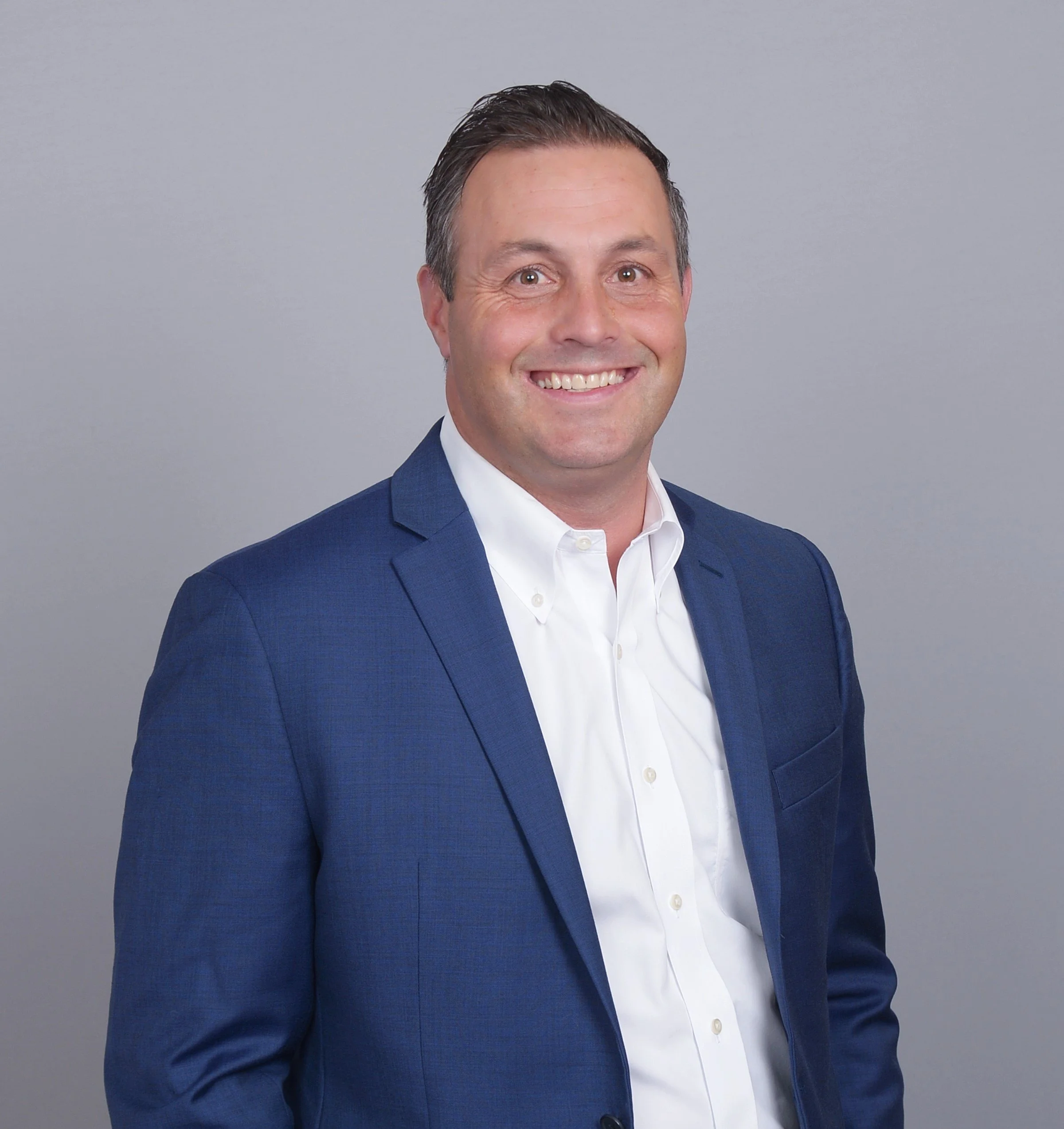 A smiling man in a blue suit and white shirt against a gray background.