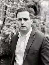 A black and white photo of a man in a suit outdoors, with trees in the background.
