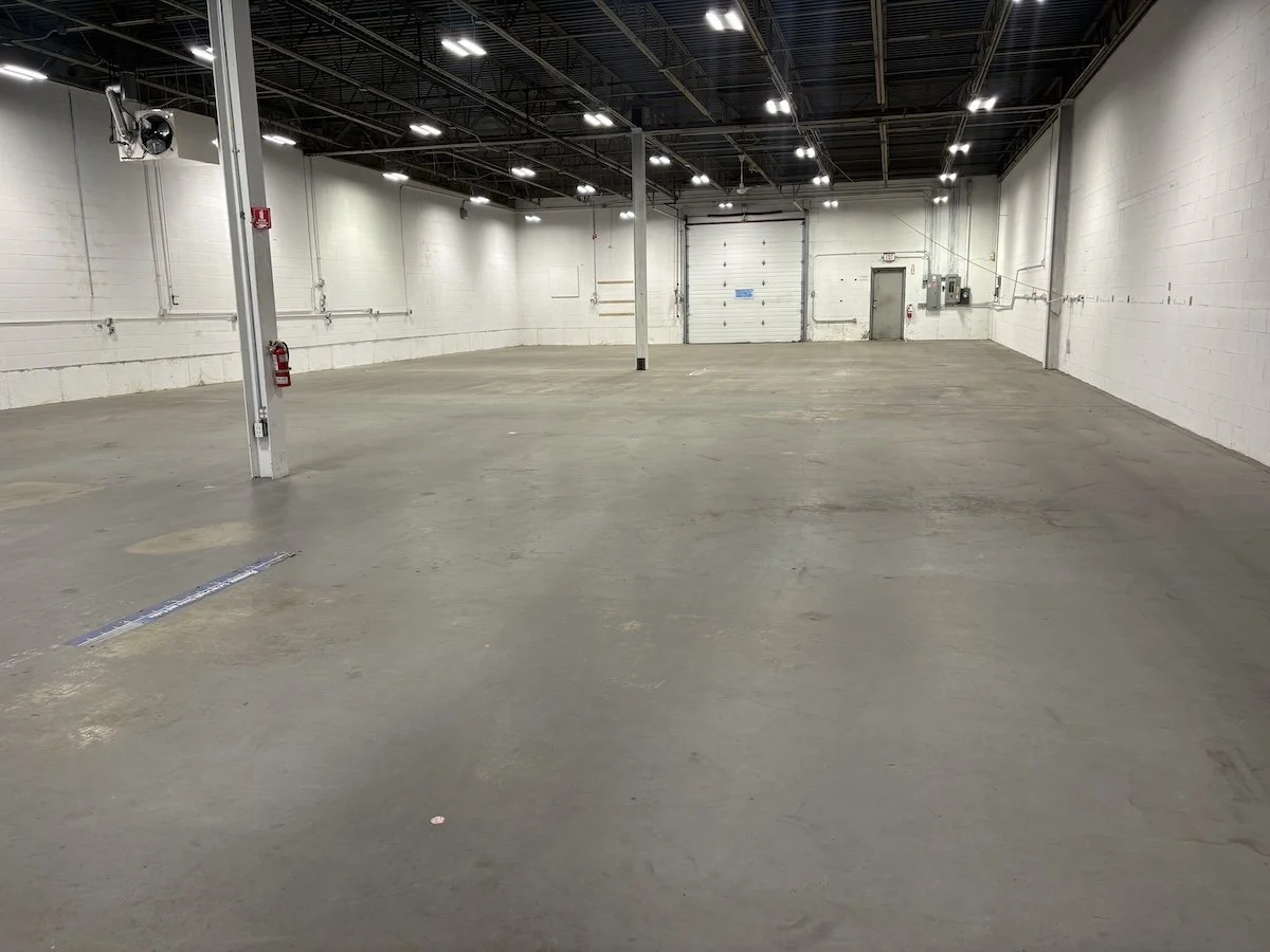 Empty indoor warehouse with concrete floor, white walls, overhead lighting, and a garage door at the back.