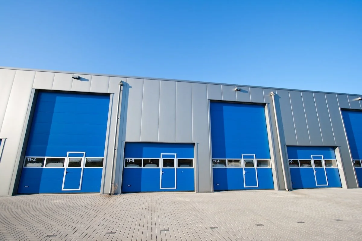 Industrial warehouse with blue rolling doors on a concrete lot under a clear blue sky.