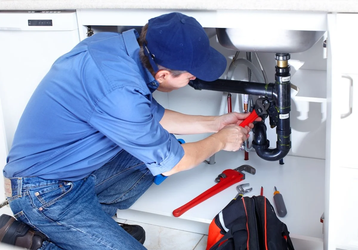 A plumber working under a kitchen sink, using a pipe wrench to tighten or loosen plumbing fittings.