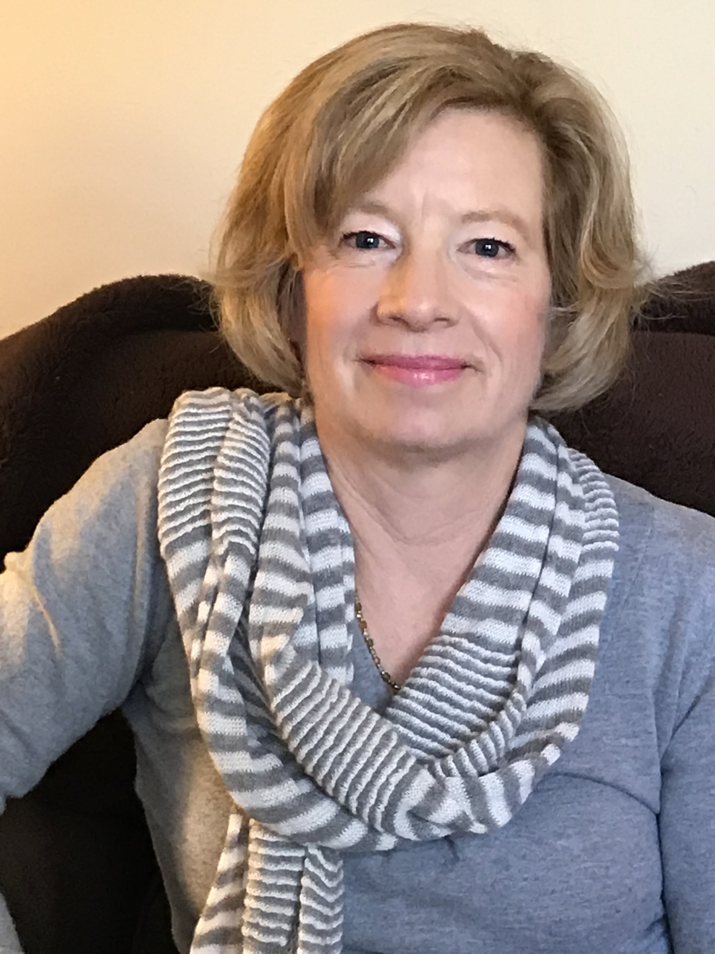 Close-up of a woman with short blonde hair, wearing a gray sweater and a striped scarf, sitting on a dark-colored piece of furniture in a room with plain walls.