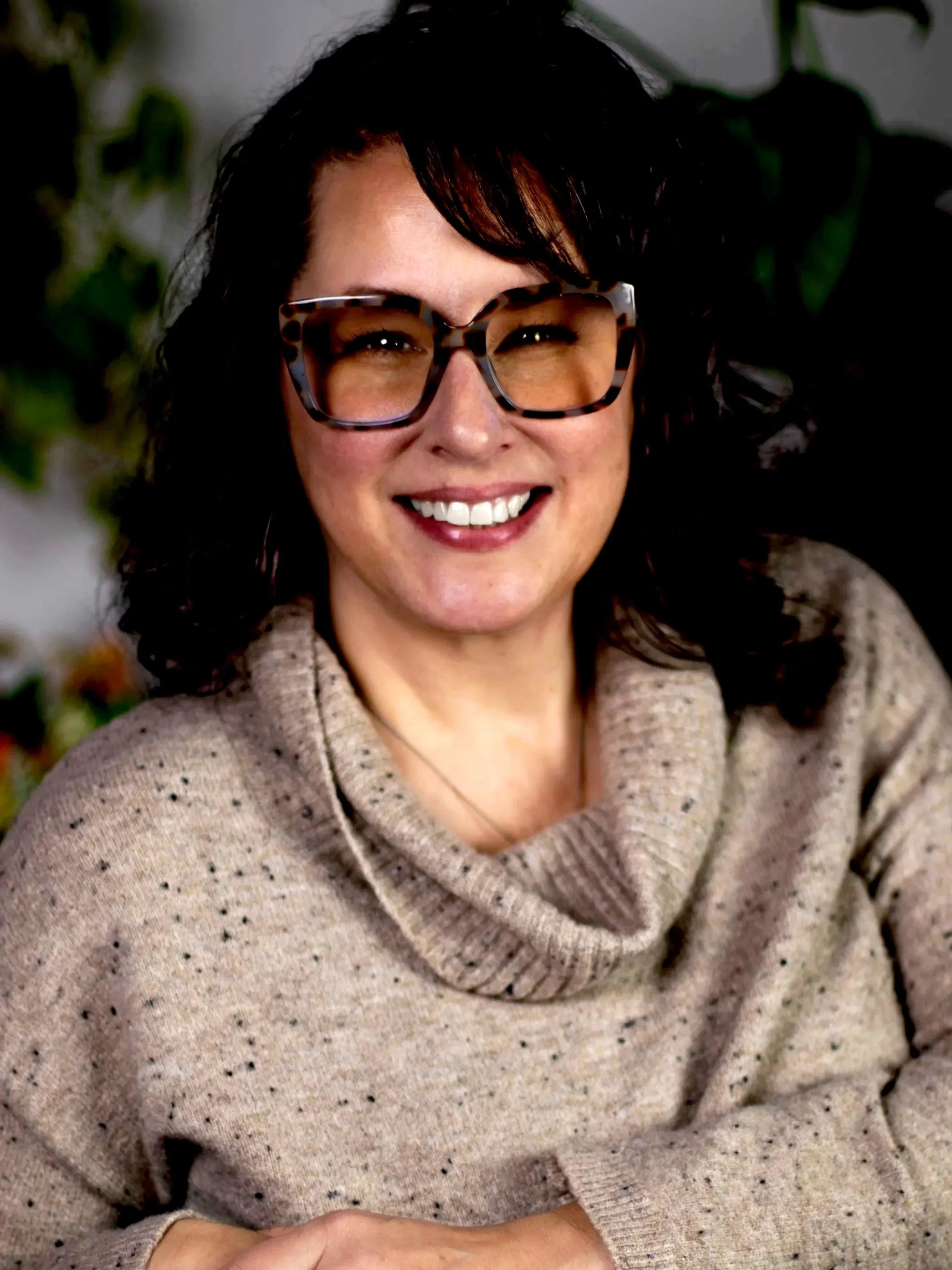 A woman with curly dark hair, wearing patterned glasses, smiling and dressed in a beige, speckled turtleneck sweater, sitting in front of a blurred background of greenery.